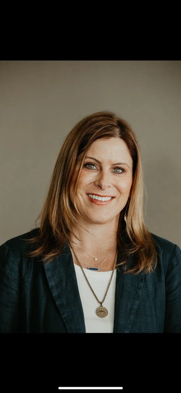 Close-up portrait of a woman with shoulder-length brown hair, blue eyes, smiling, wearing a dark blazer, a necklace with a round pendant, and a white shirt against a neutral background.