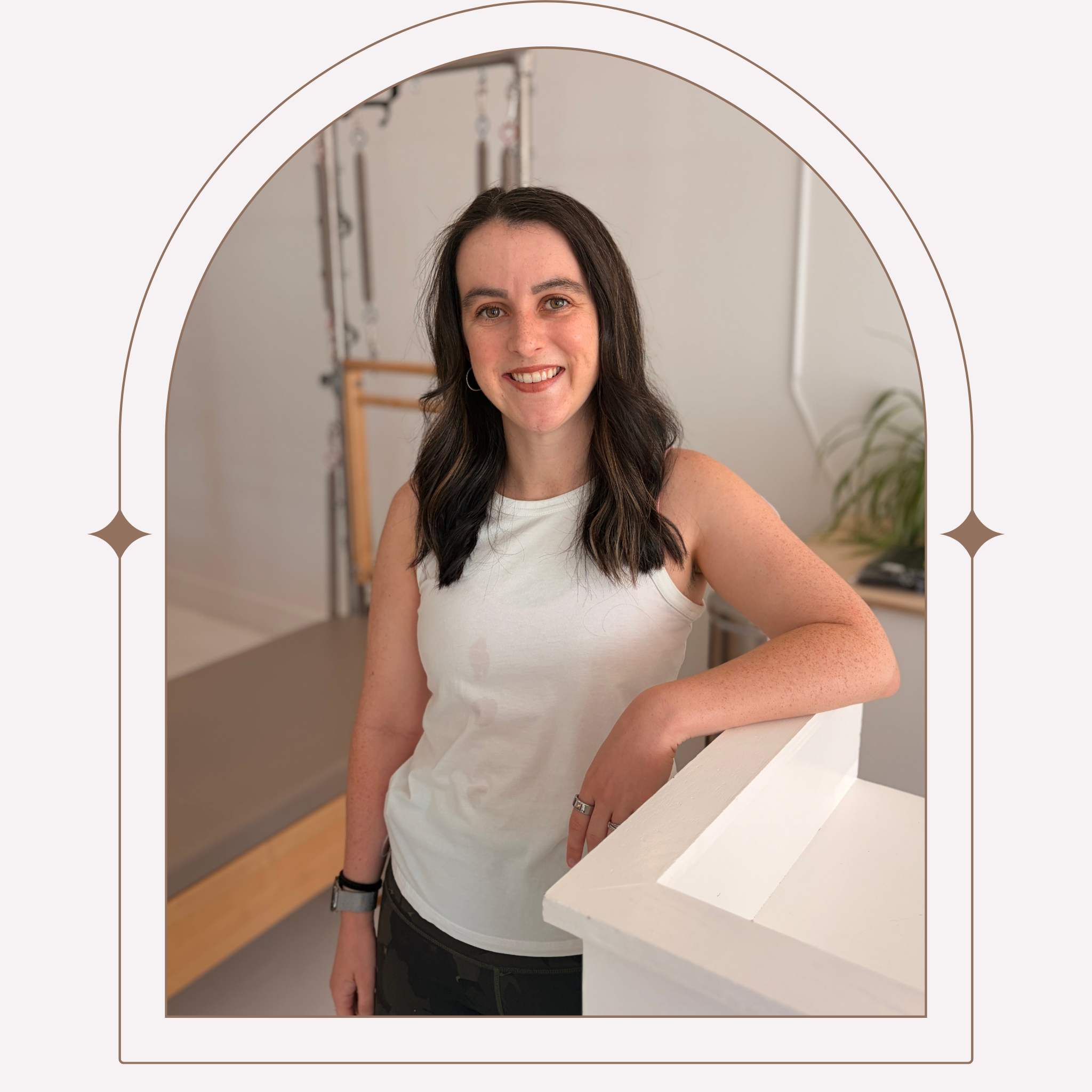 A woman with black hair wearing a white t-shirt and a smartwatch, smiling and leaning on a white counter in a room with white curtains and a small wooden table in the background.