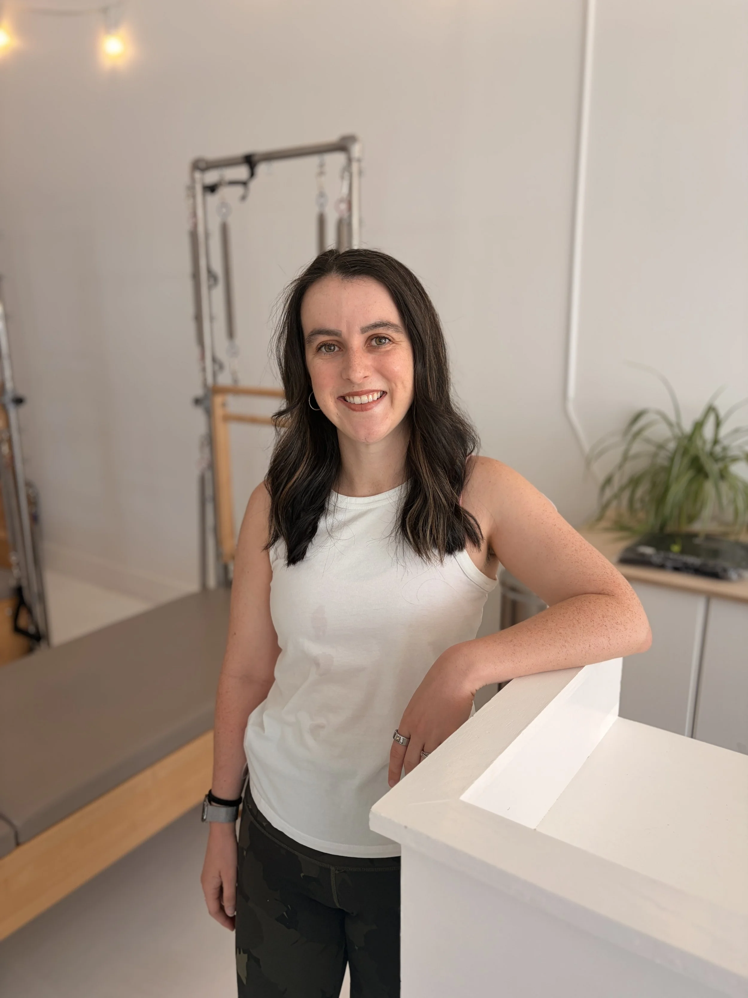 A woman with black hair, smiling, wearing a white t-shirt, standing in front of a white background.