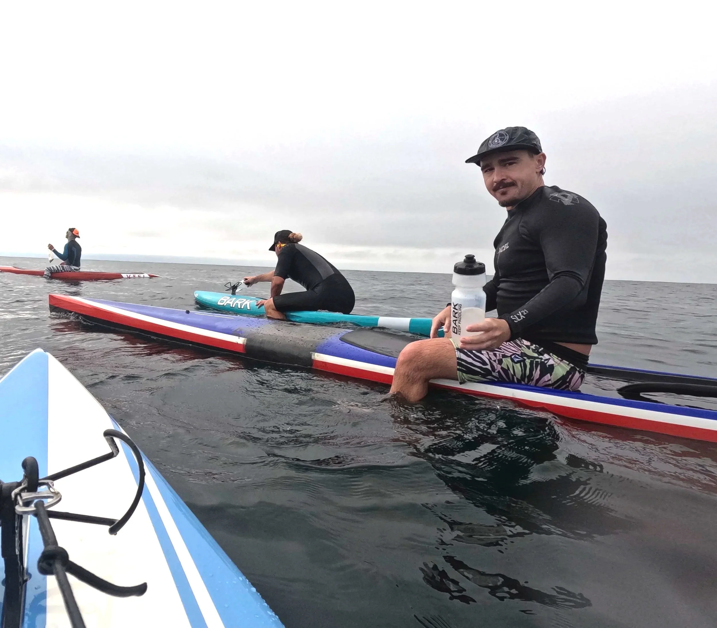 A man lying on a paddleboard on calm water, looking at the camera, with another person paddleboarding in the distance.