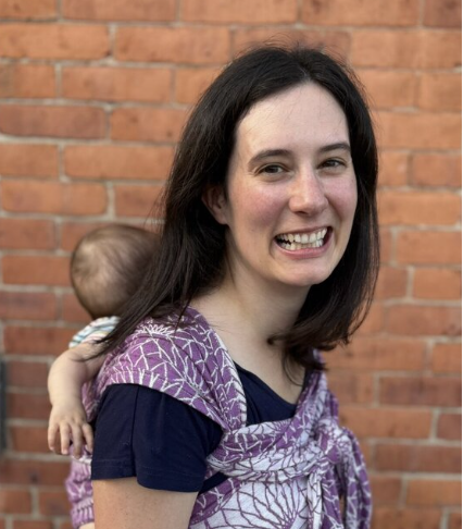 Woman smiling with a baby's head resting on her shoulder, standing in front of a brick wall.