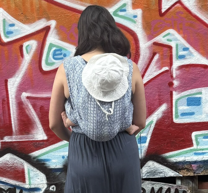 A woman with dark hair wearing a blue patterned sleeveless top and black pants, standing with her back to the camera in front of a colorful graffiti wall. She has a white hat hanging from her backpack.