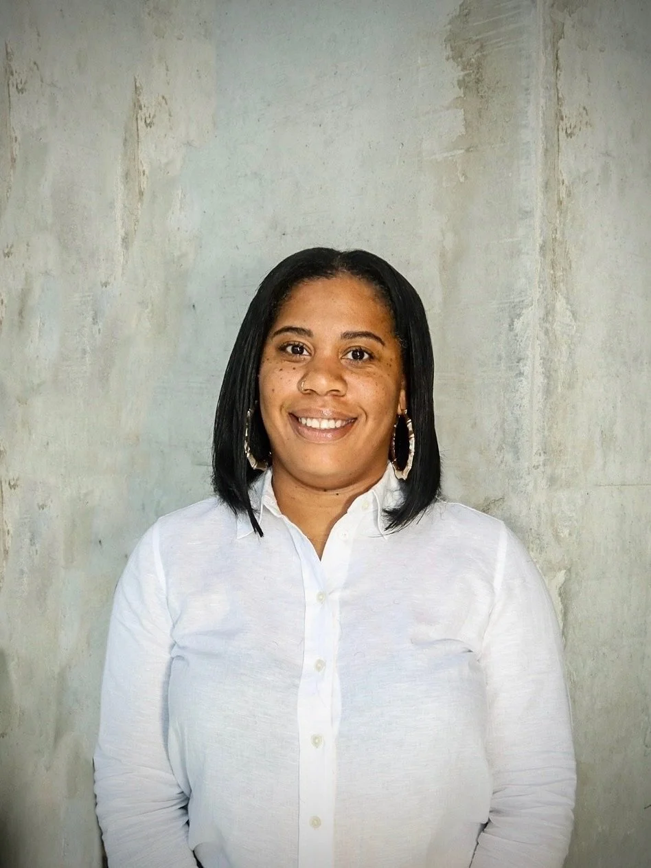 A woman with shoulder-length black hair, wearing a white button-up shirt and hoop earrings, smiling in front of a textured gray wall.