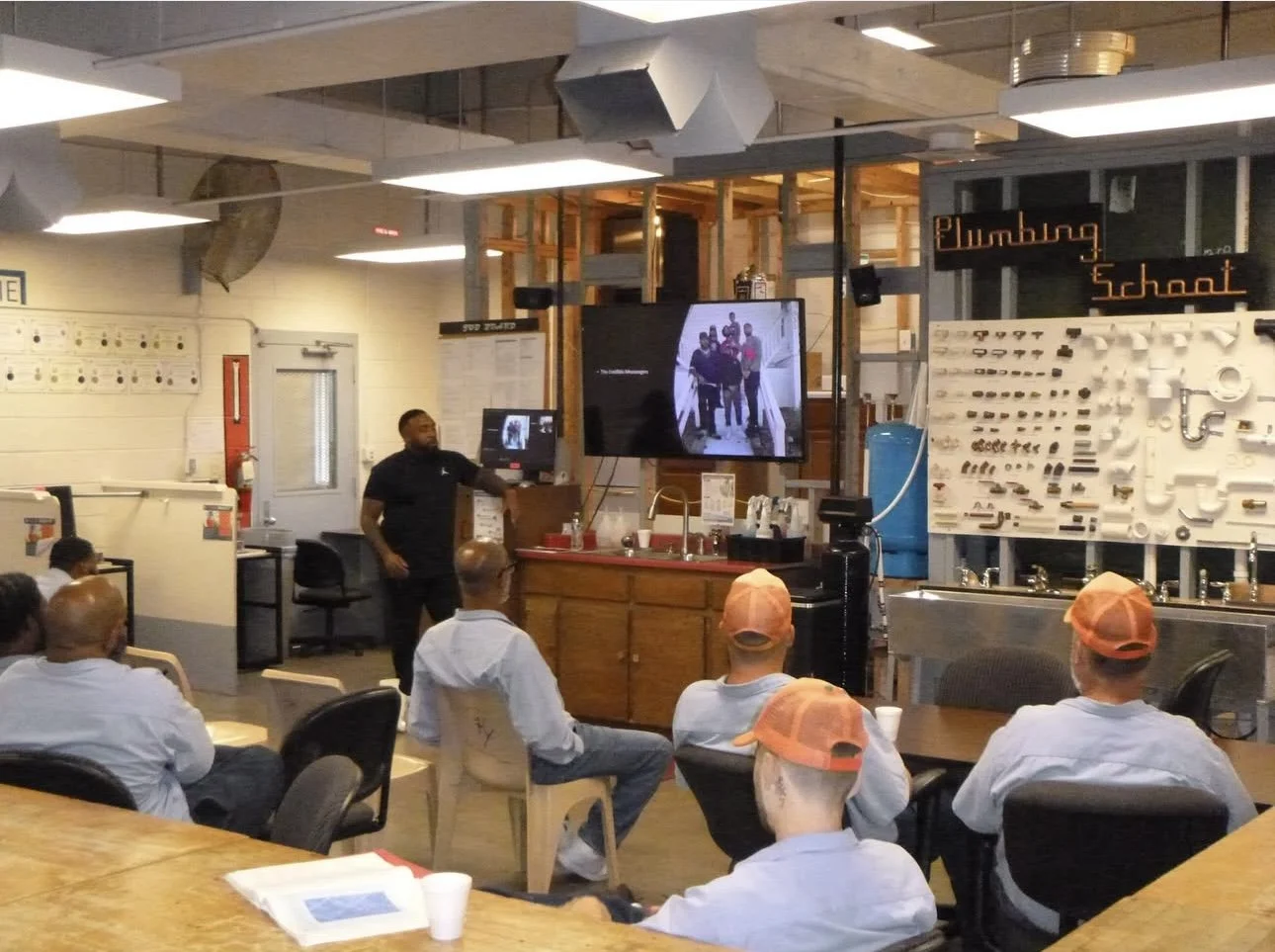 A diverse group of young people participating in a discussion or group activity around a table in a classroom, with a whiteboard in the background containing handwritten notes and a message with names.