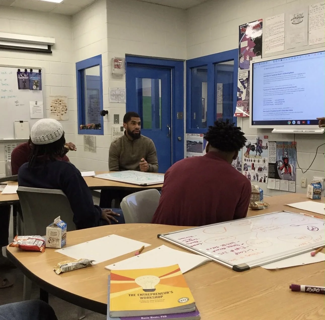 A group of young people in a classroom or meeting room sitting in a circle, with some standing and speaking. The room has large windows, a whiteboard, and some tables and shelves in the background.