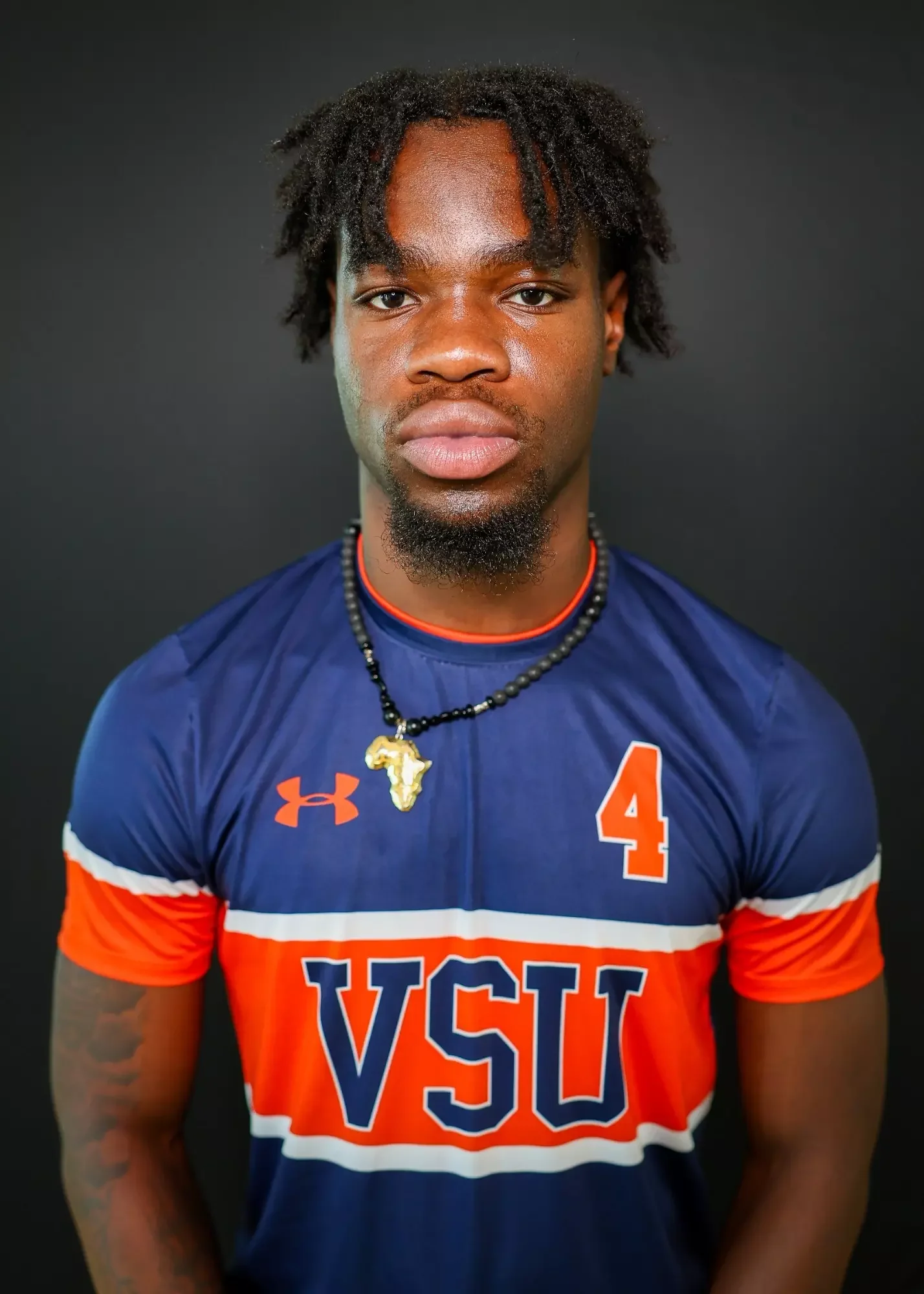 Portrait of a young man wearing a blue and orange sports jersey with VSU and the number 4, and a necklace with an Africa-shaped pendant, standing against a black background.