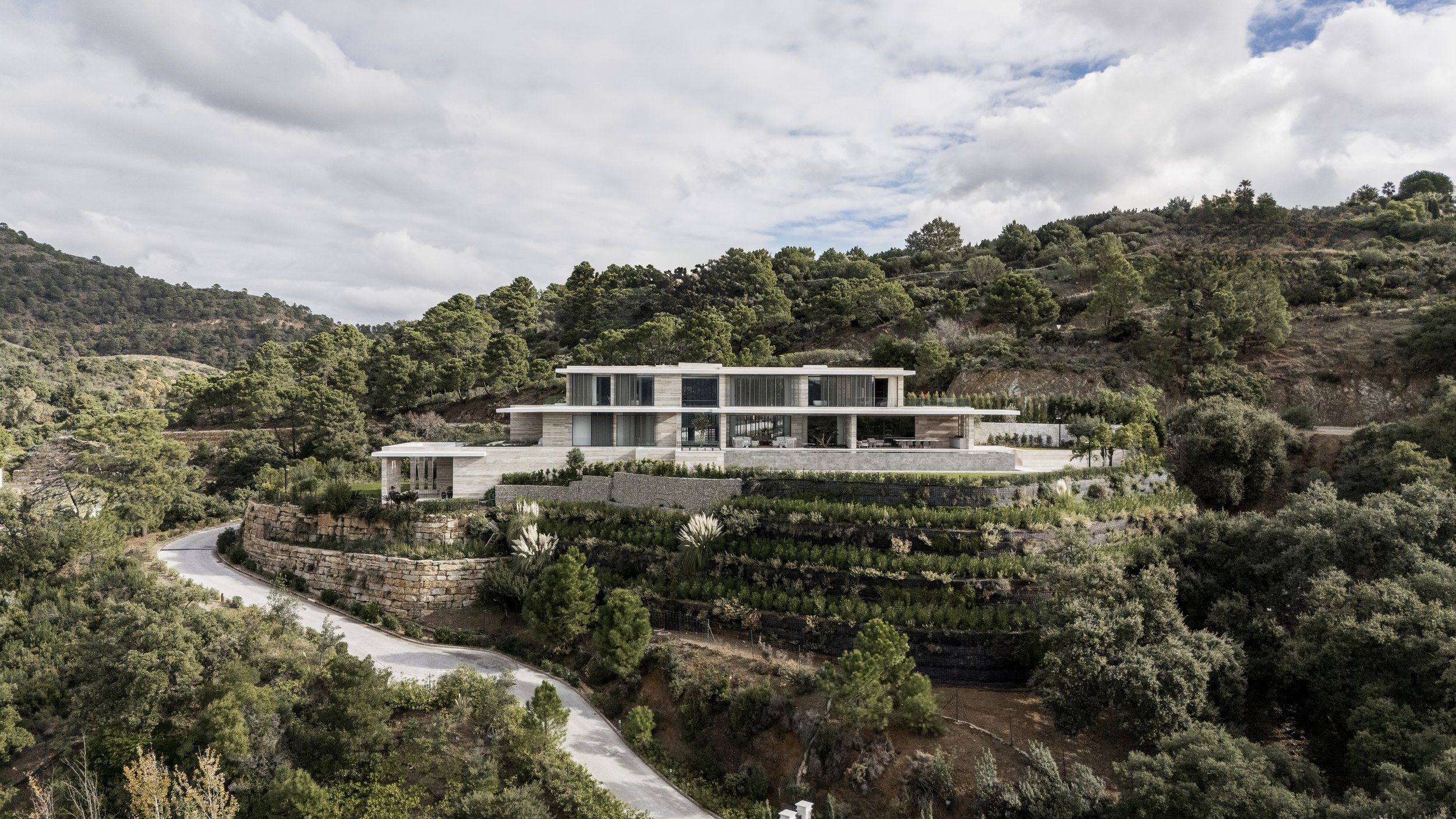Casa moderna de varios niveles en una colina, rodeada de árboles y vegetación, con camino curvado en primer plano.
