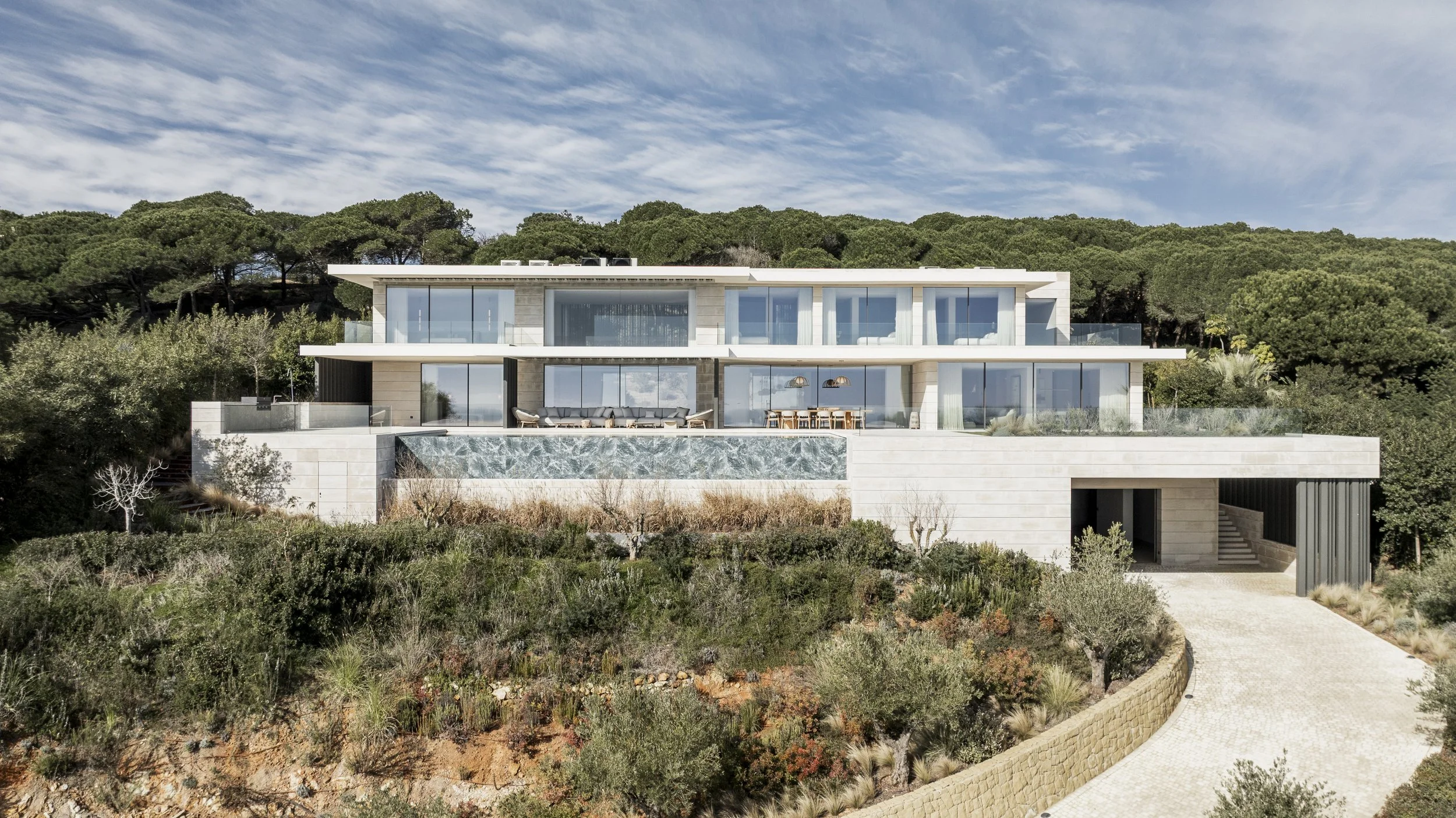 Casa moderna de varias plantas en un entorno natural con árboles y una pequeña colina, con grandes ventanas y un camino de entrada de piedra.