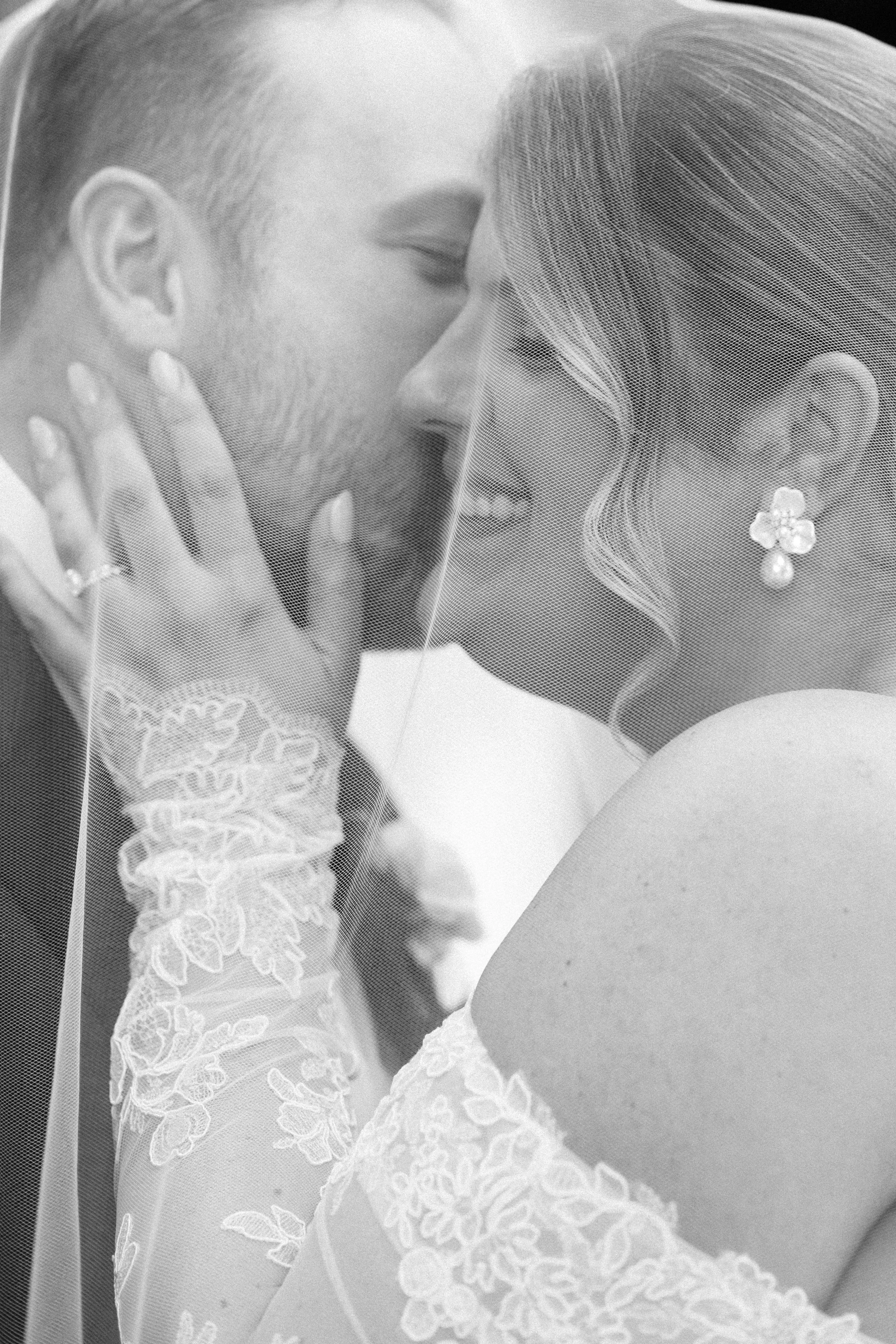 A close up photograph of a bride and groom underneath a veil while the groom kisses her cheek.