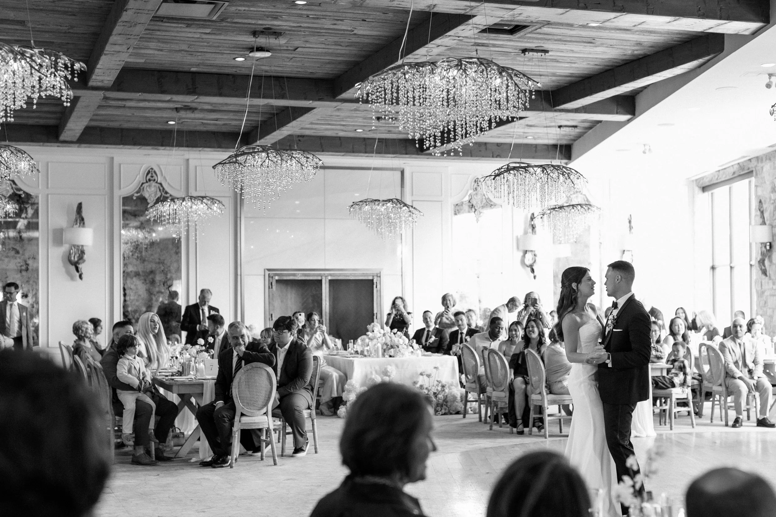 A black and white photo of a wedding reception with a bride and groom dancing in the center, surrounded by seated guests at tables, in a decorated banquet hall with chandeliers and large windows.