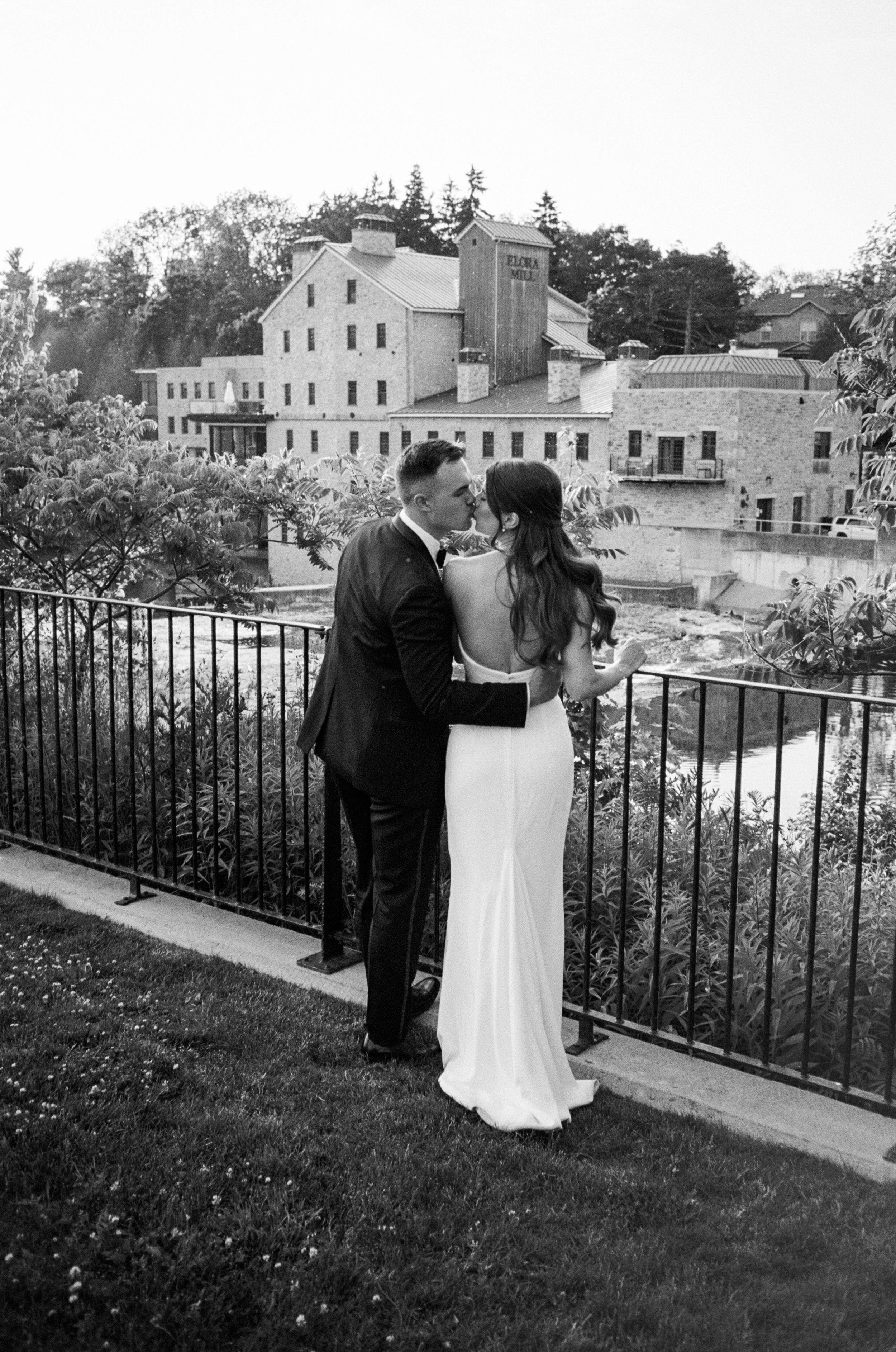 A newlywed couple kissing while facing the famous Elora Mill across the river in black and white film.