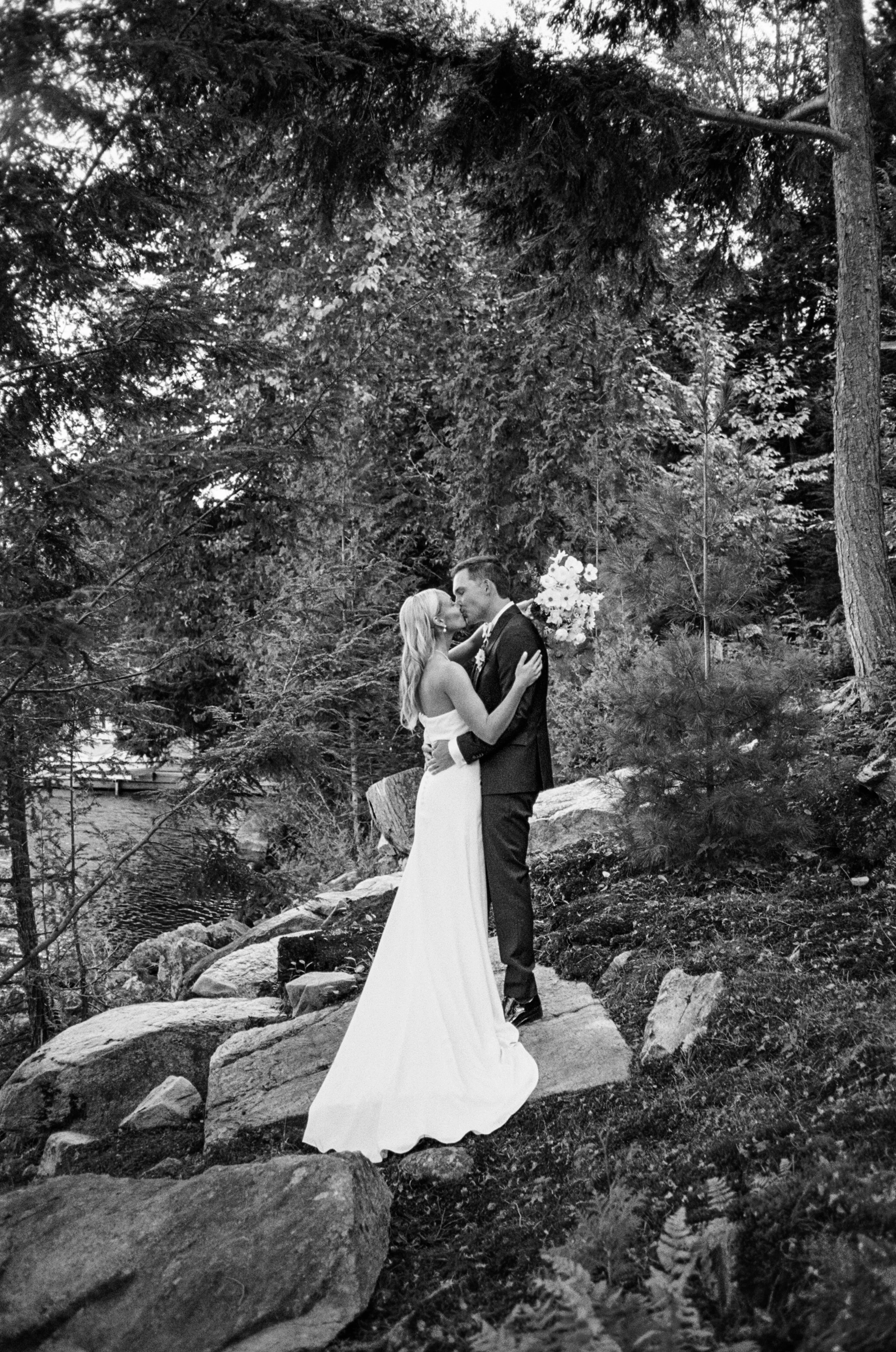 A black and white photo of a bride and groom standing on Muskoka rock amongst the greenery of their cottage in Muskoka, Ontario.