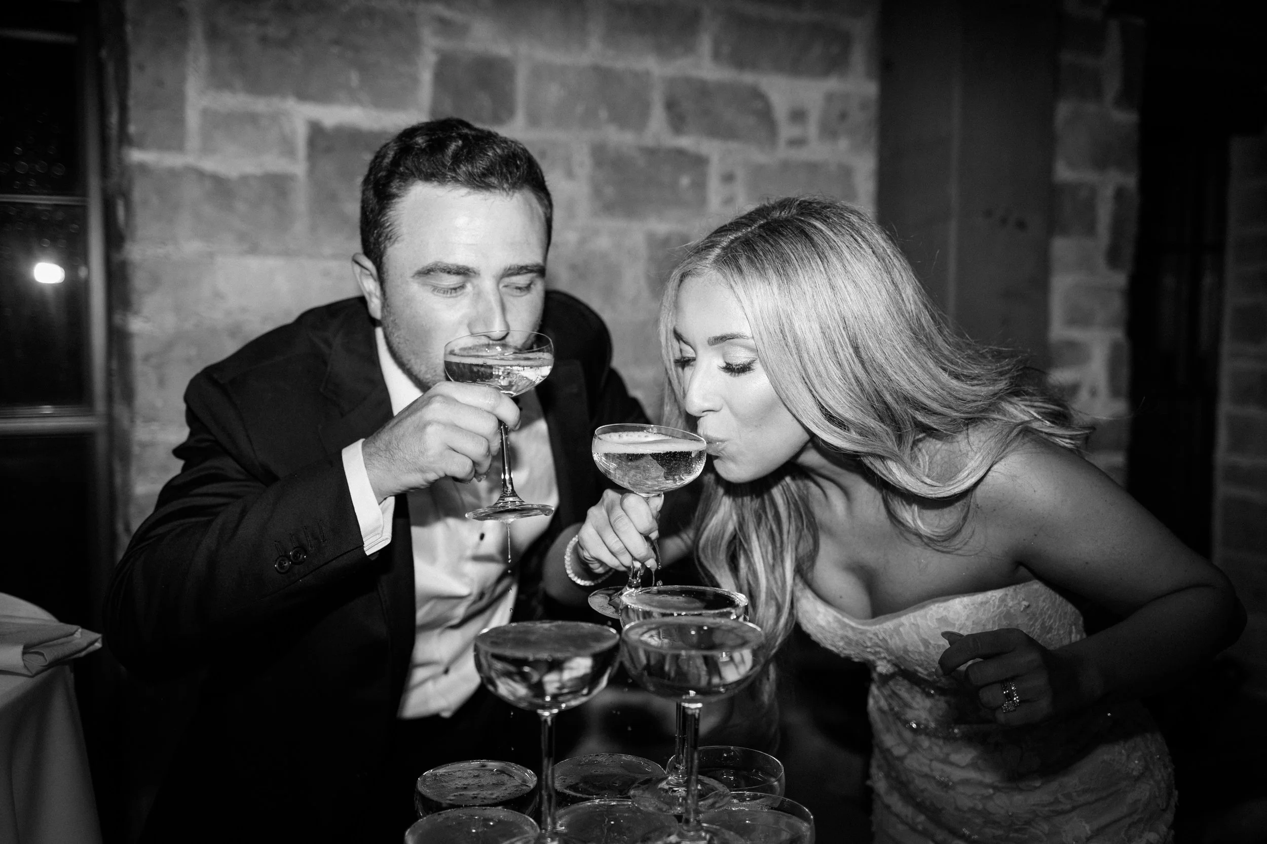 A man and woman in formal attire are drinking champagne from coupe glasses, toasting at a celebration event.