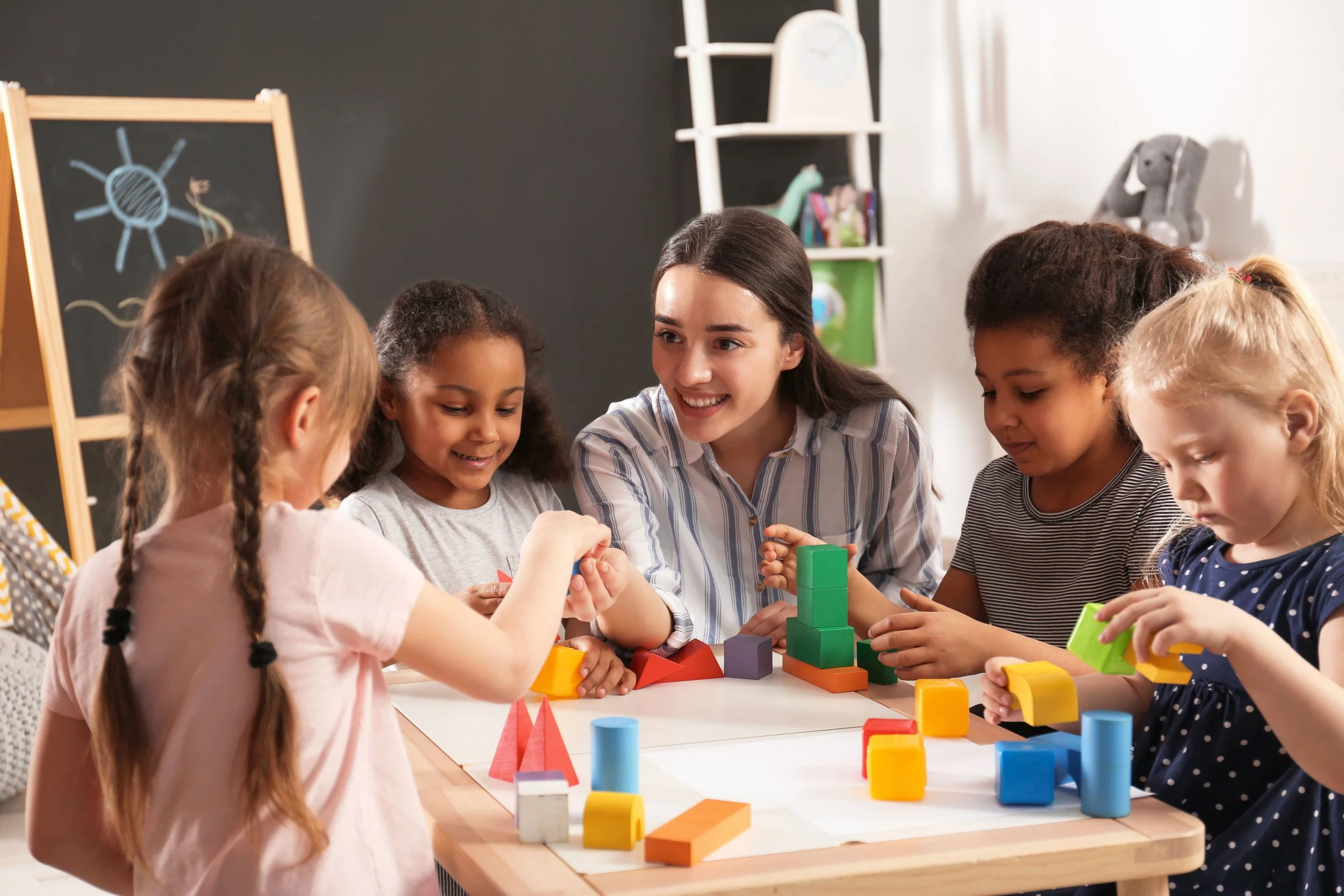 A young woman teaching a group of five young girls how to play with colorful building blocks at a table in a classroom.