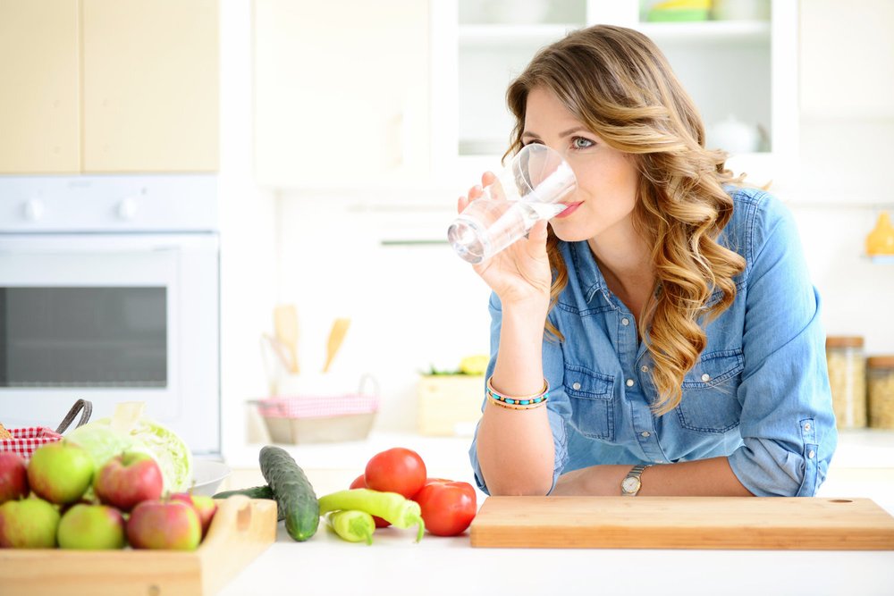 A woman drinking a glass of water at a kitchen counter with fresh fruit and vegetables