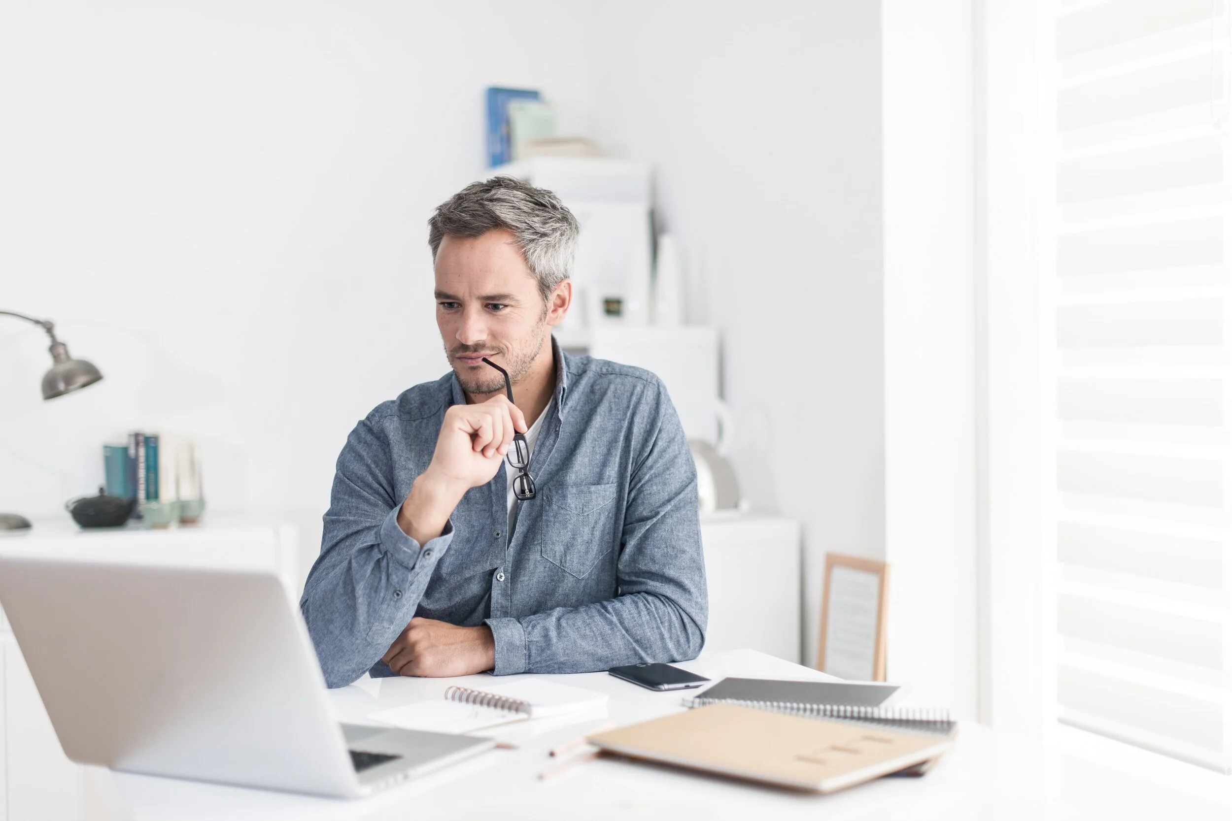 A man sitting at a white desk, looking at a laptop, holding glasses near his chin, with a thoughtful expression, in a bright office space.