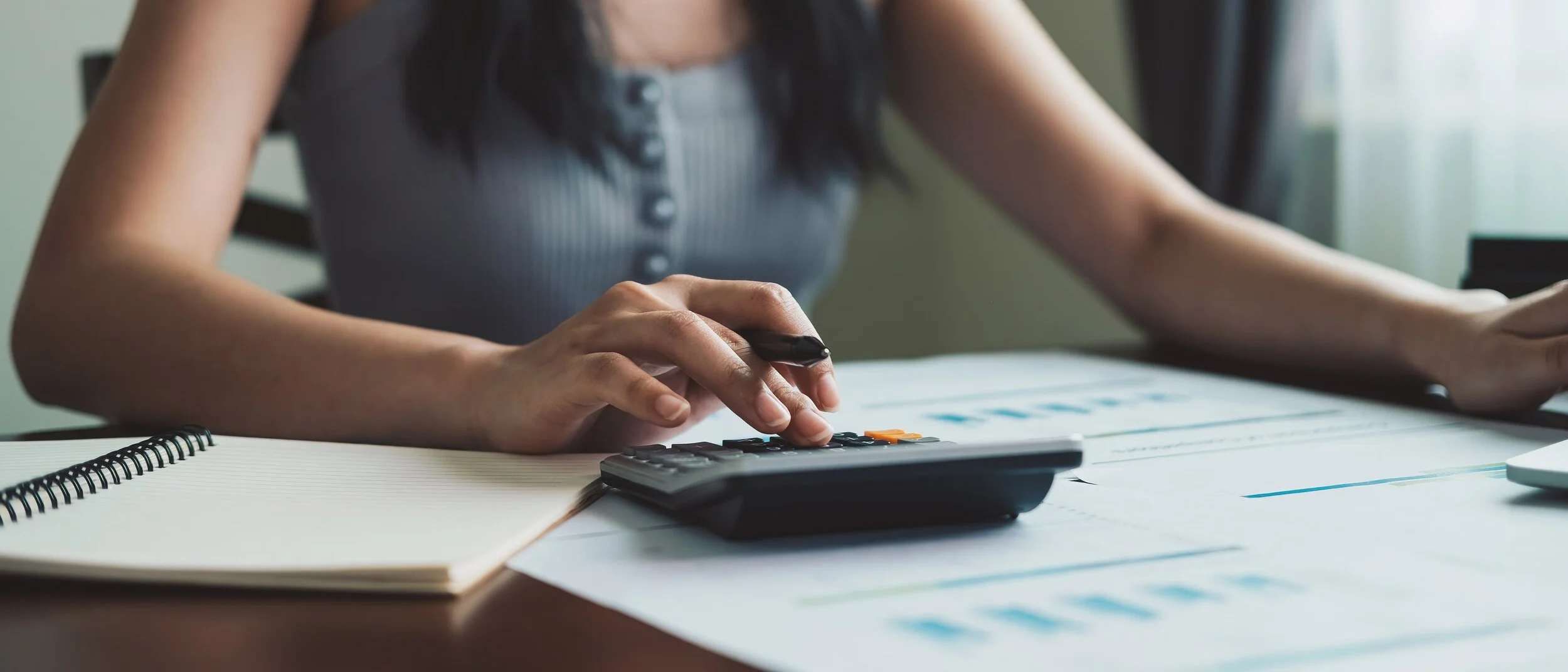 A woman working at a desk with a calculator, notebook, and printed charts, possibly analyzing financial data.