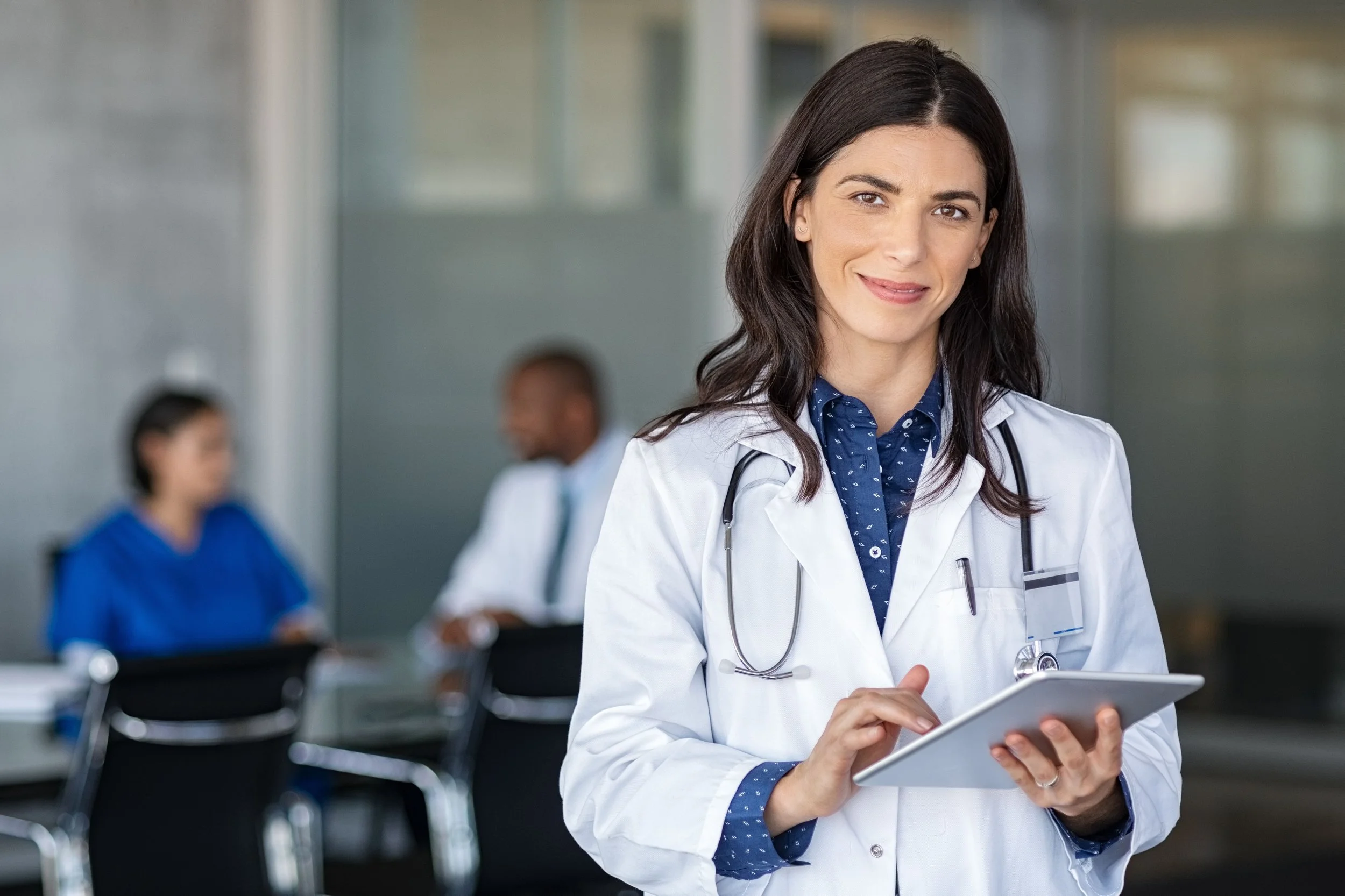 Female doctor with stethoscope and tablet in a hospital or medical office setting, with blurred colleagues in the background.