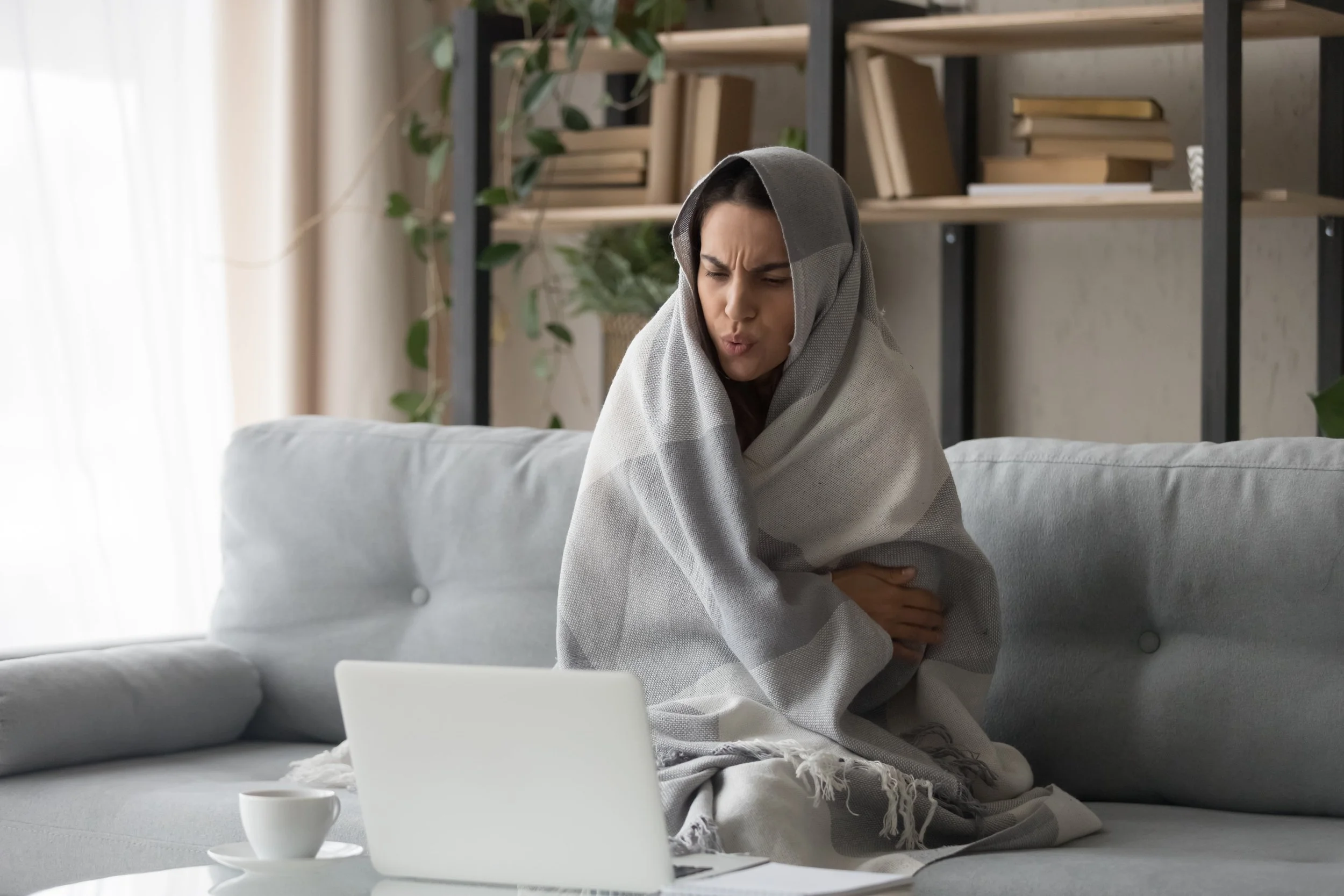 A woman in her living room, cold wrapped in a blanket using her laptop computer