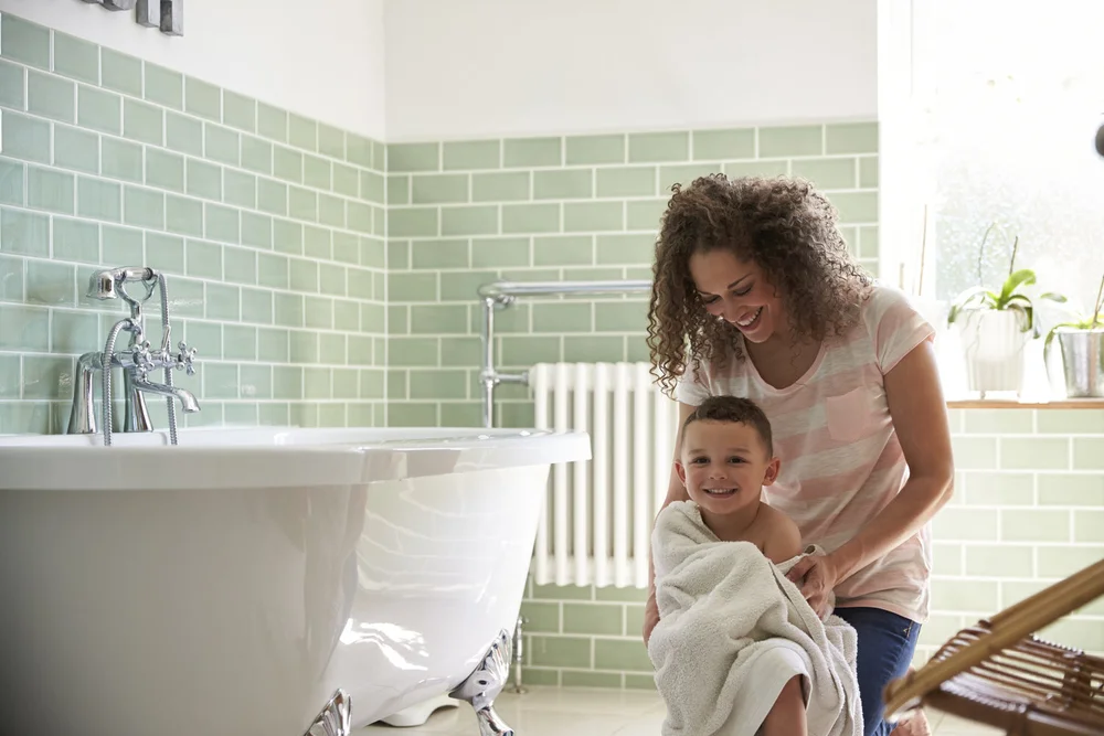 A mother wrapping her smiling young son in a towel after his bath, beside a white clawfoot tub