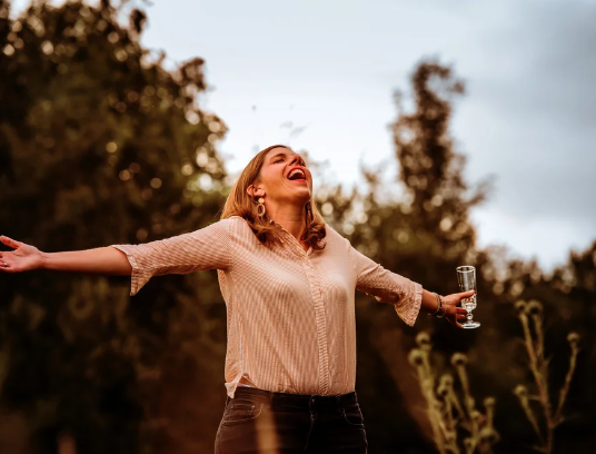 Vrouw met open armen en glas champagne, glimlachend in een natuurlijke omgeving