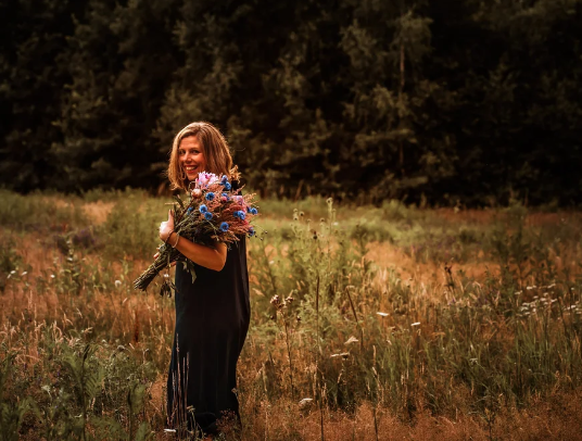 Vrouw met een bloemenboeket in een weiland, omgeven door gras en struiken, met een bos op de achtergrond.