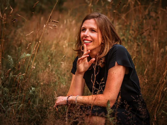 Vrouw die in een veld zit en lacht, omgeven door gras en planten.