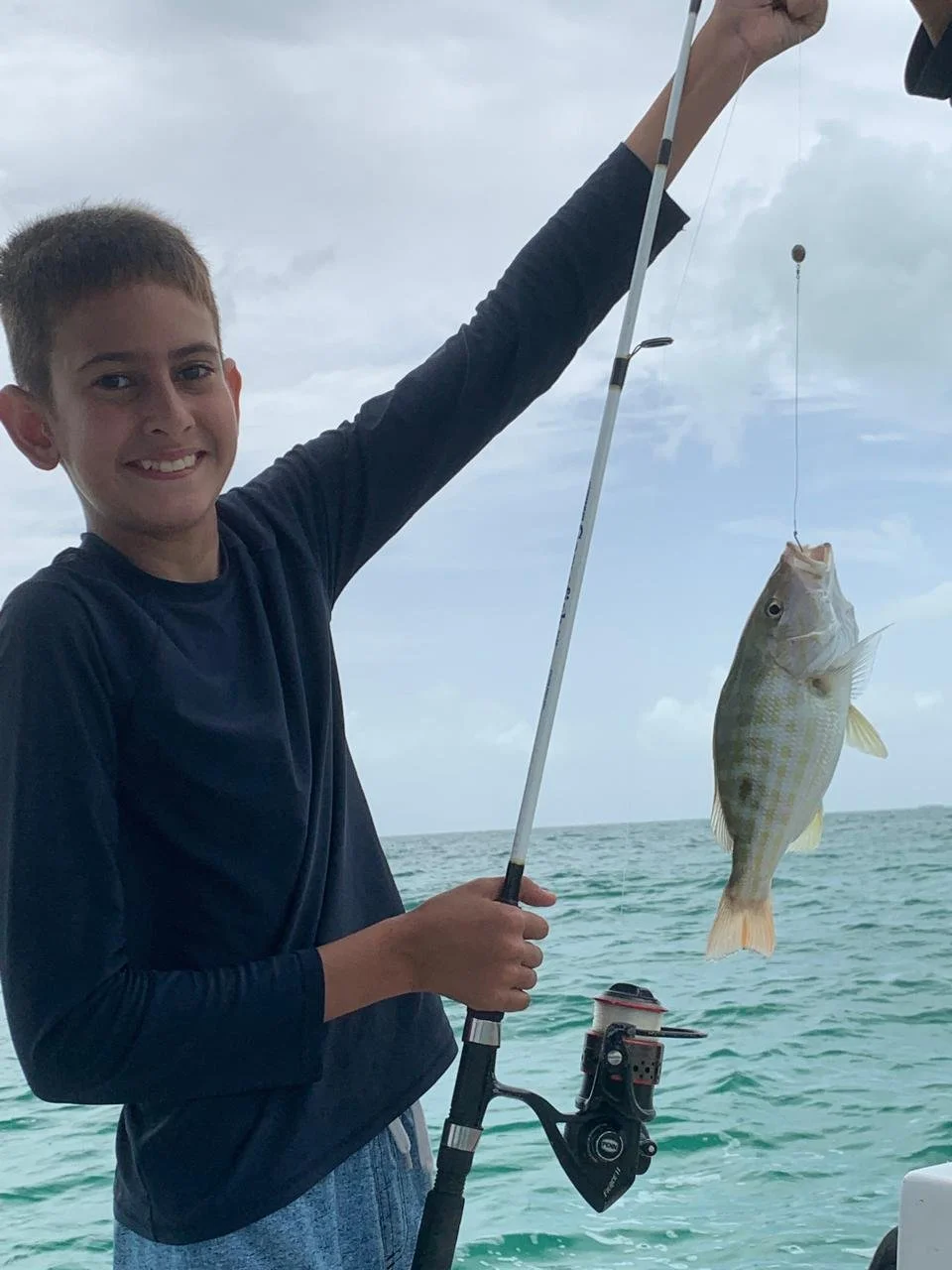 A smiling boy on a boat in San Pedro Belize holding up a fishing rod with a fish he just caught.