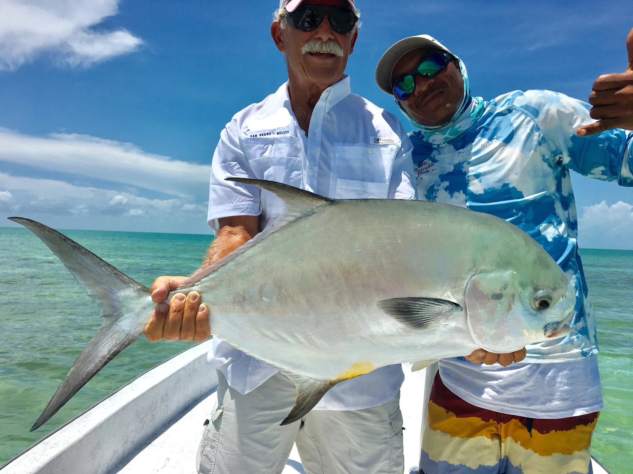 An angler Ken Watkins and a local fishing guide smiling on a boat holding a large permit fish caught on the turquoise flats near San Pedro, Ambergris Caye, Belize.