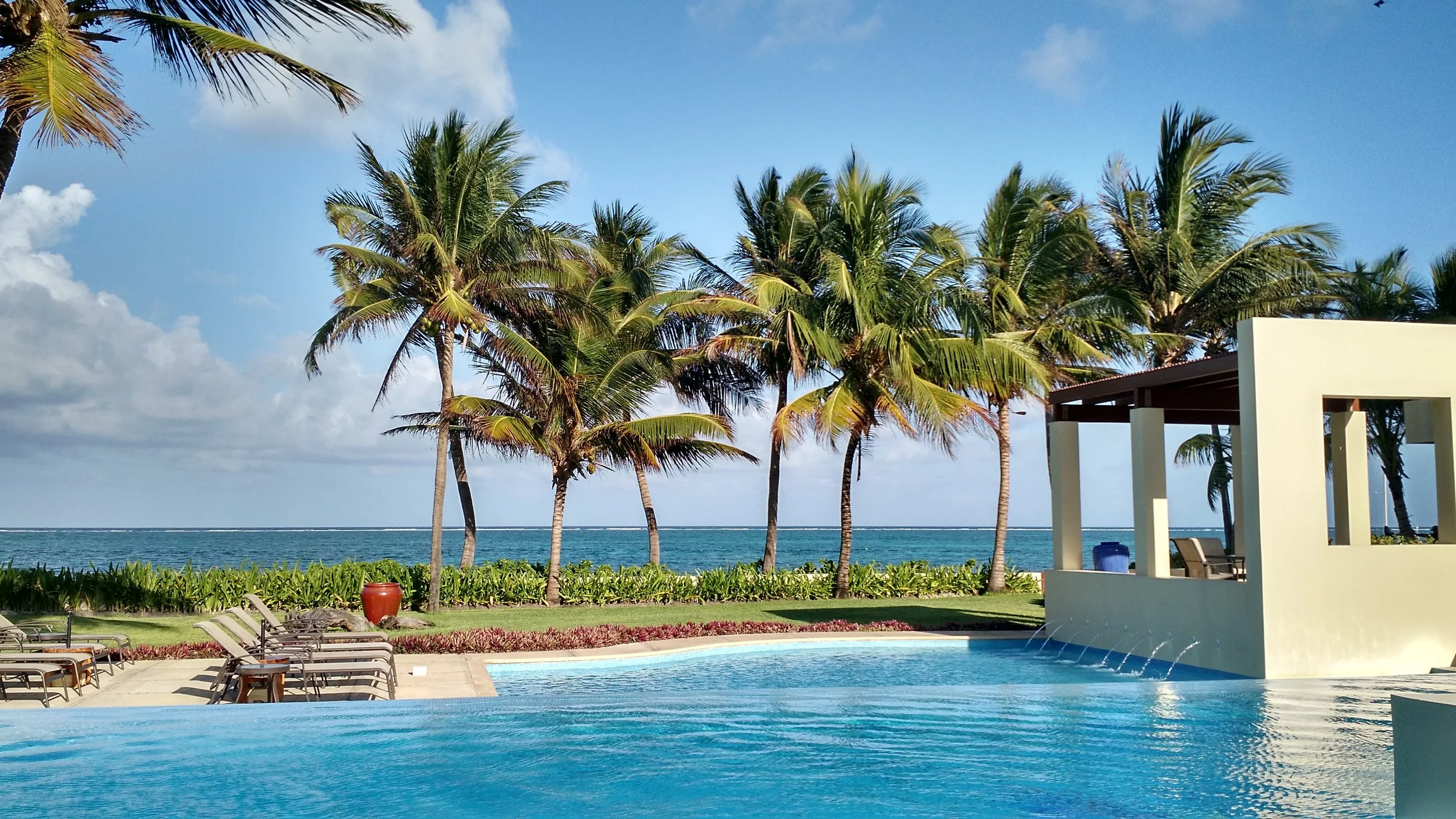 Sunlit oceanfront swimming pool at The Phoenix Resort surrounded by lush green lawns, lounge chairs, and tall coconut palm trees overlooking the Caribbean Sea in San Pedro, Belize.