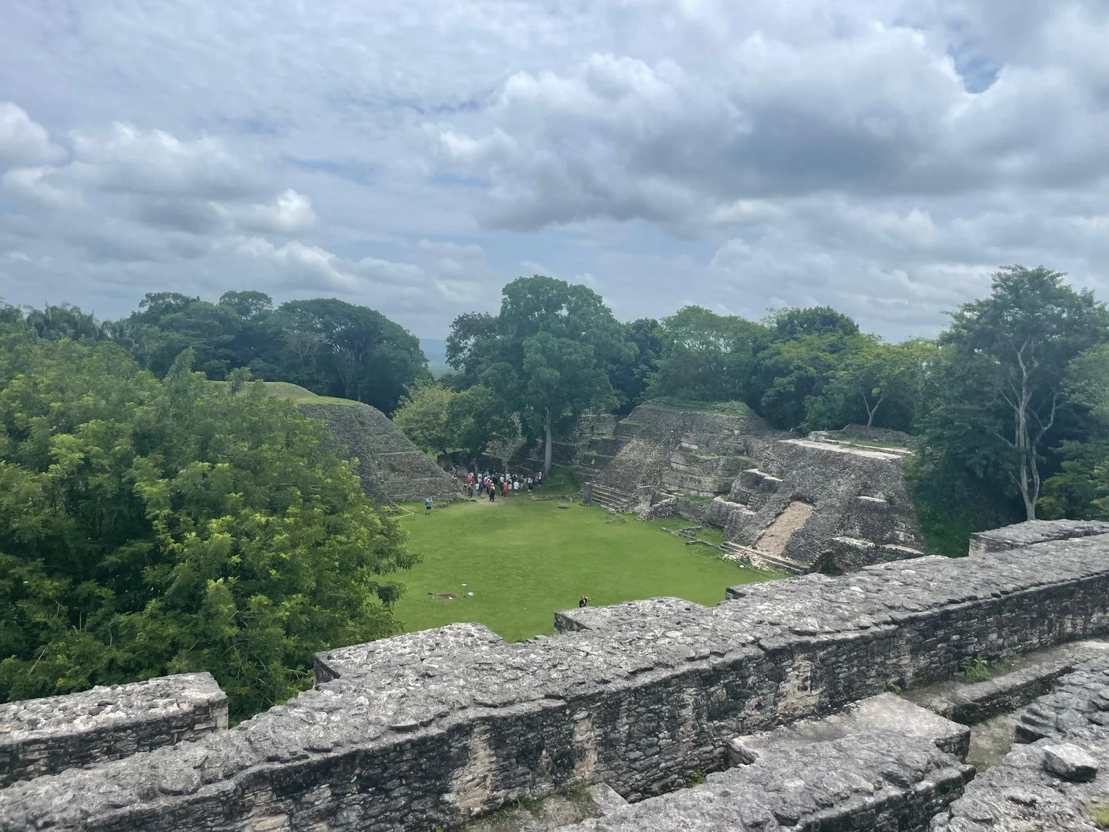 High angle view of a Mayan plaza surrounded by lush green jungle with a small group of tourists below.
