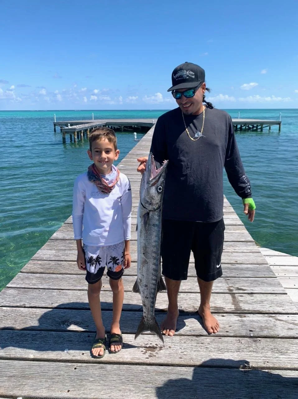 A young guest and a local guide standing on a wooden dock at The Phoenix Resort in San Pedro, Belize, holding up a large barracuda they caught.
