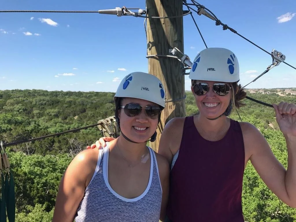 Two women wearing safety helmets and sunglasses smiling before a zip lining tour over the tropical canopy in mainland Belize.