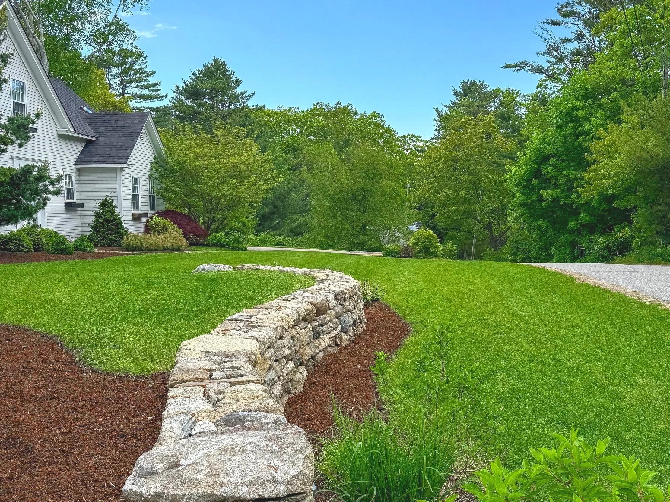 A well-maintained front yard with a stone retaining wall, green lawn, and a white house surrounded by trees under a partly cloudy sky.