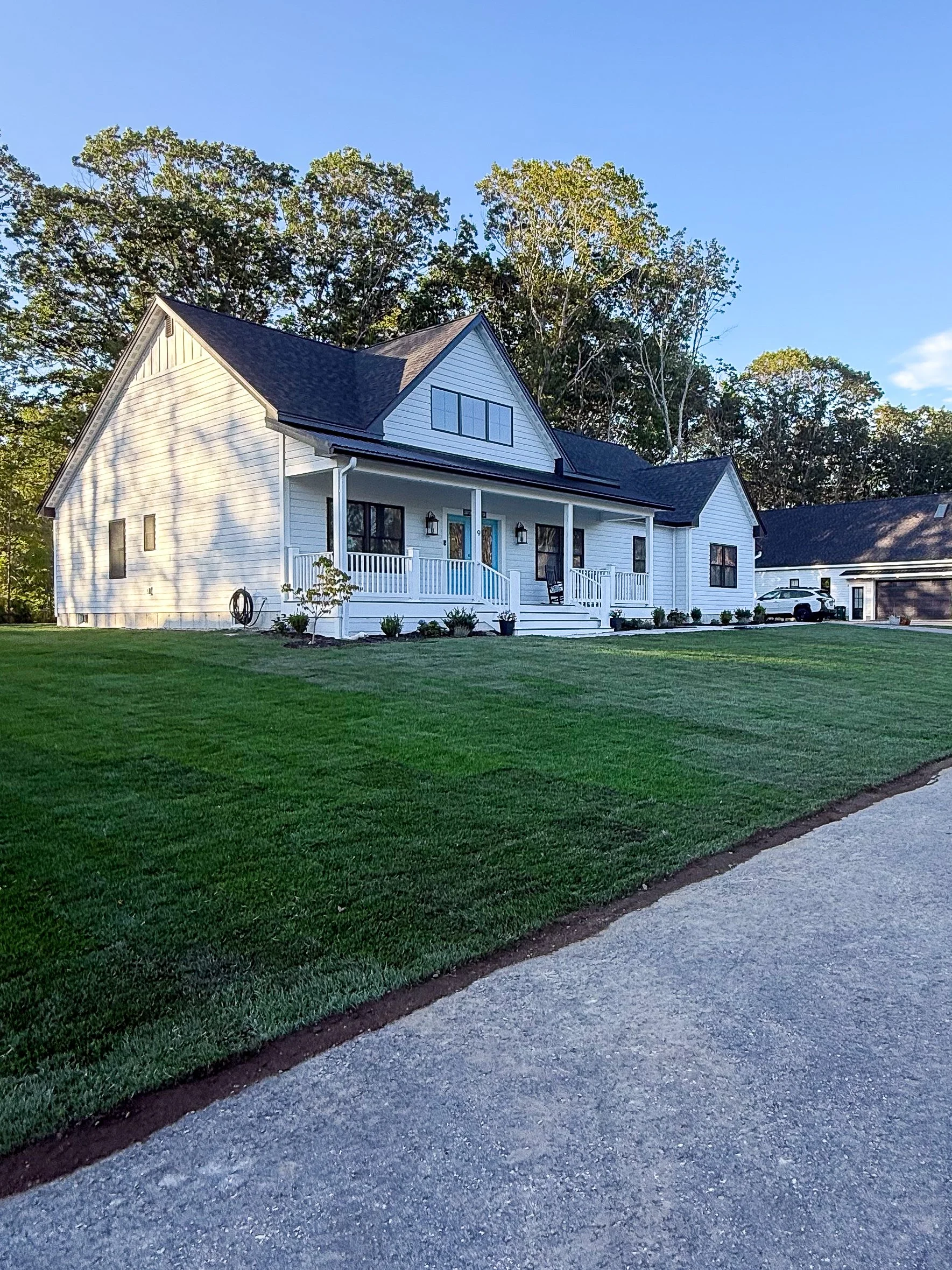 A white two-story house with a front porch, gabled roof, black shutters, and surrounded by a well-maintained lawn and trees in the background.