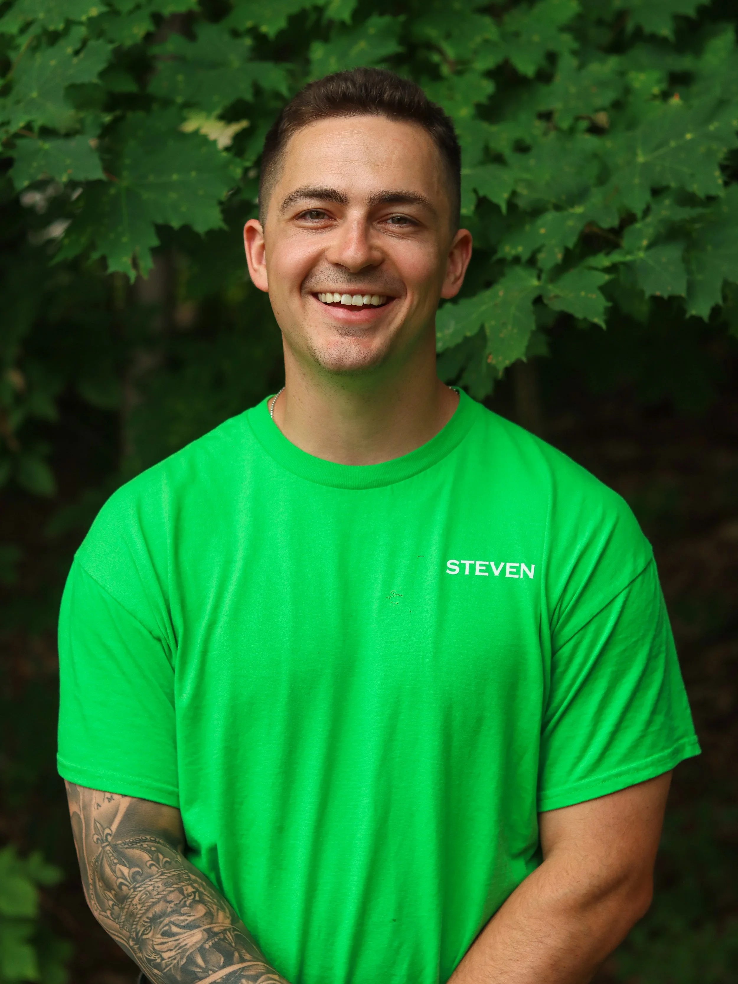 Man with green shirt and dark hair smiling with trees and forest behind him.