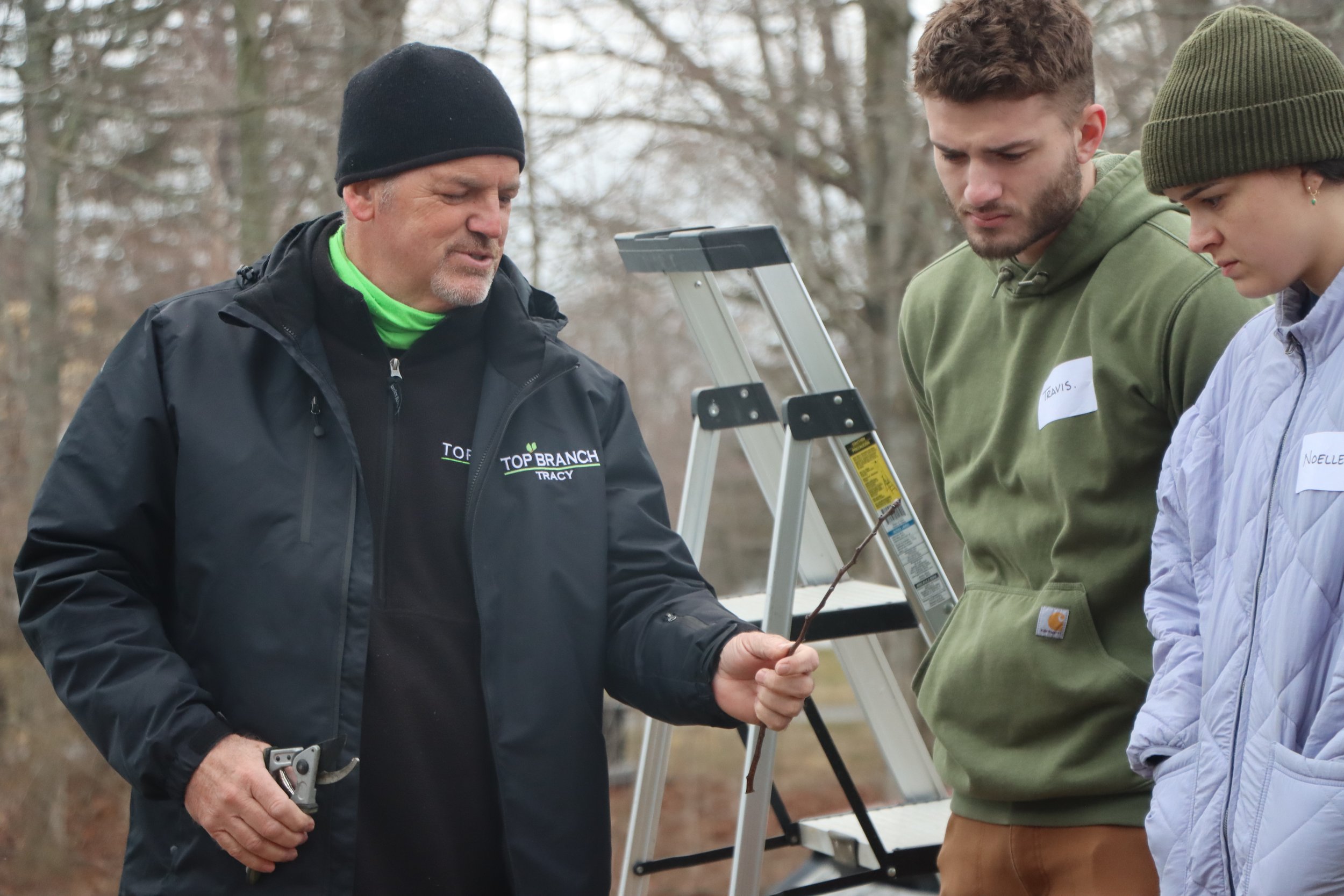 Tracy Chaplin of Top Branch Tree Service one man demonstrating and explaining how to prune apple trees and inspected buds on a stick to a younger man and woman, near a ladder, trees in the background, overcast sky.