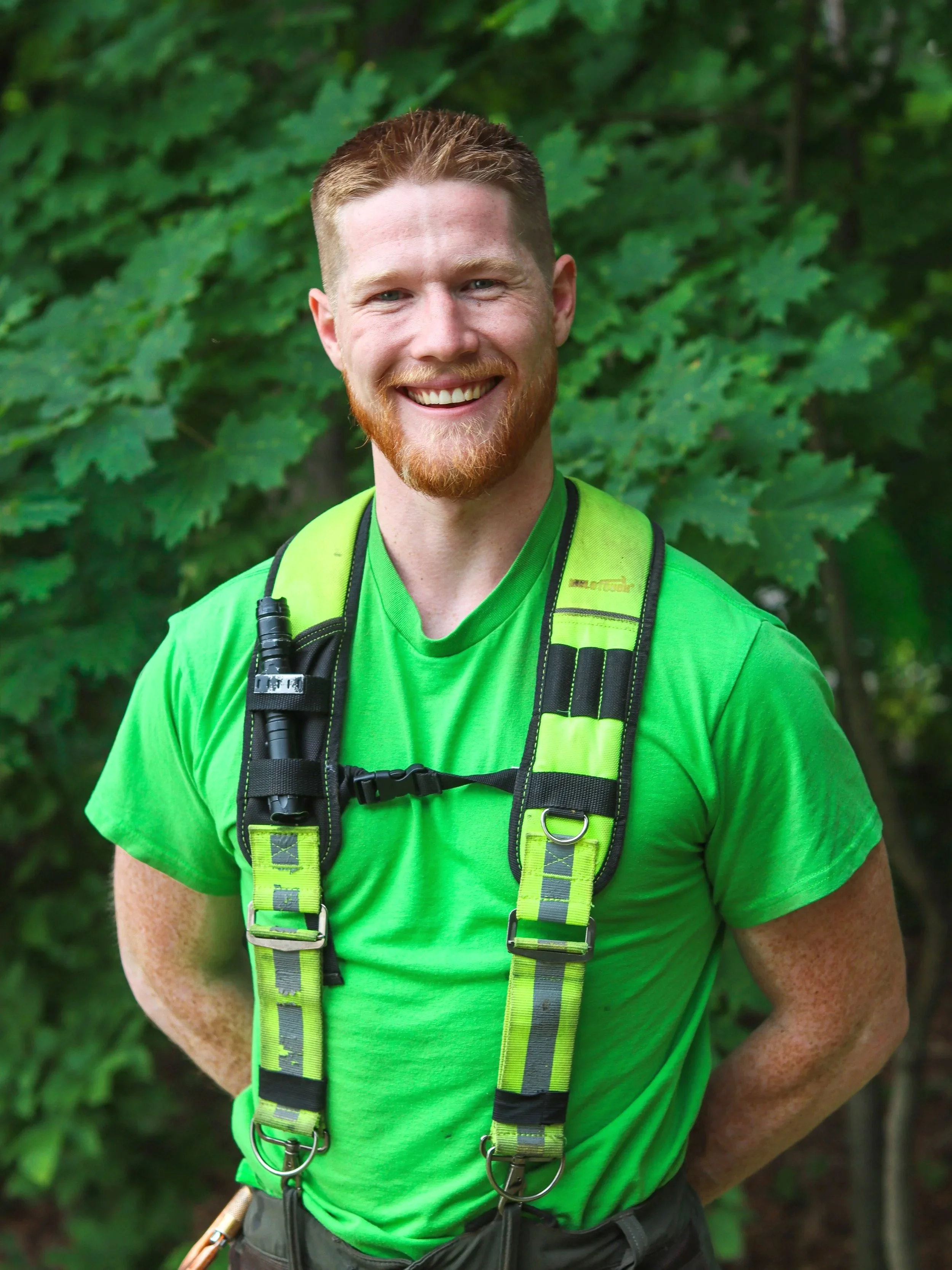 A man with a beard wearing a green shirt takes a selfie outdoors. Behind him is a small white house with a blue door and windows, surrounded by trees and a green lawn. A black truck is parked near the house.