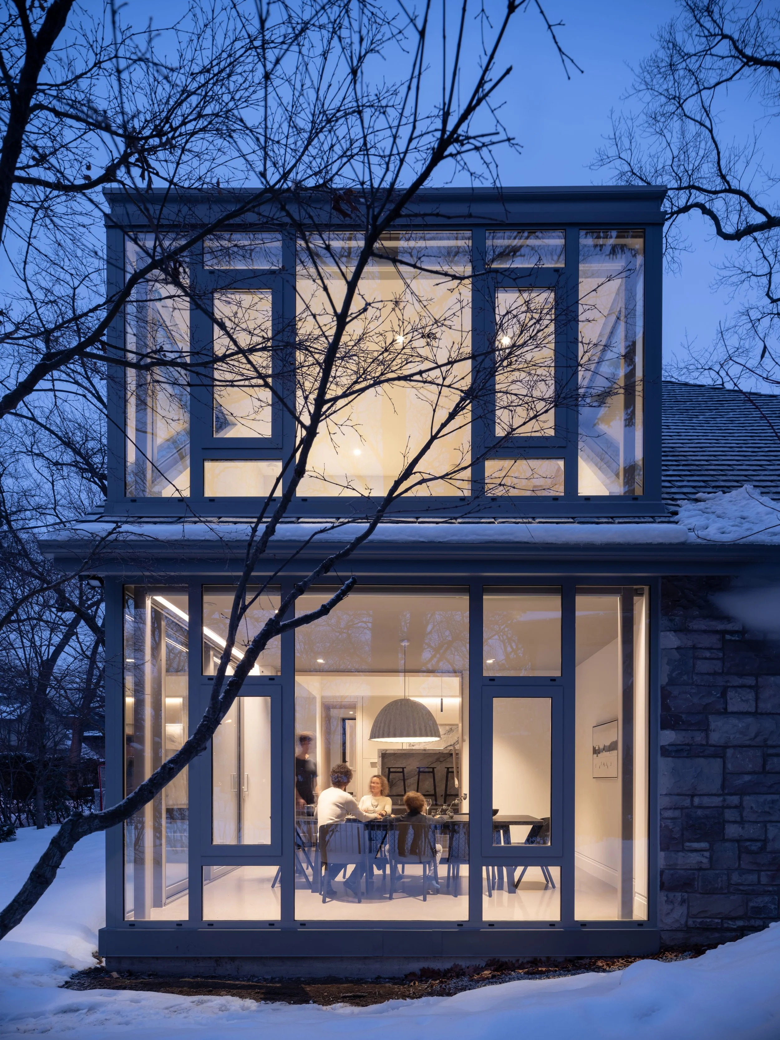 A cozy indoor gathering seen through large glass windows of a house at dusk, with snow outside and several people sitting around a table, socializing.
