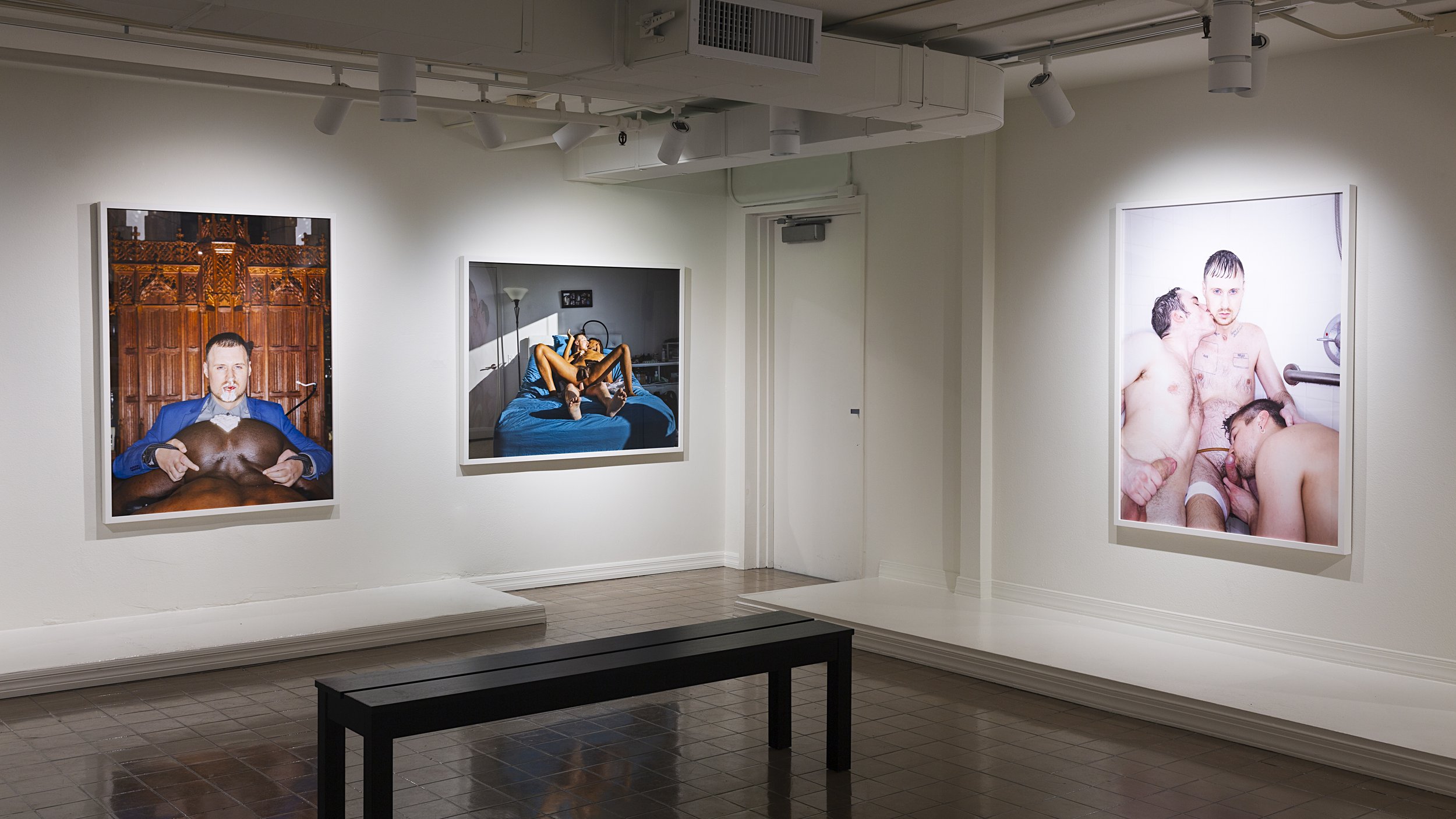 Gallery with large photographs on the walls, featuring intimate and provocative scenes. A bench is in the foreground.