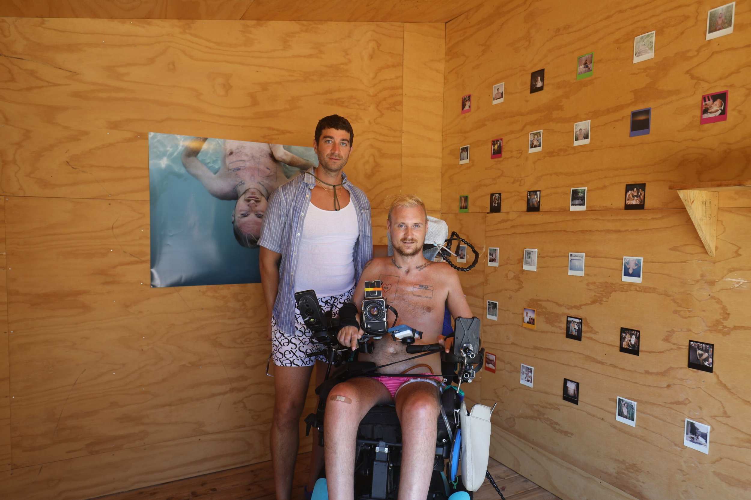 Two people inside a wooden room, one person standing and the other in a power wheelchair, surrounded by photos on the walls.