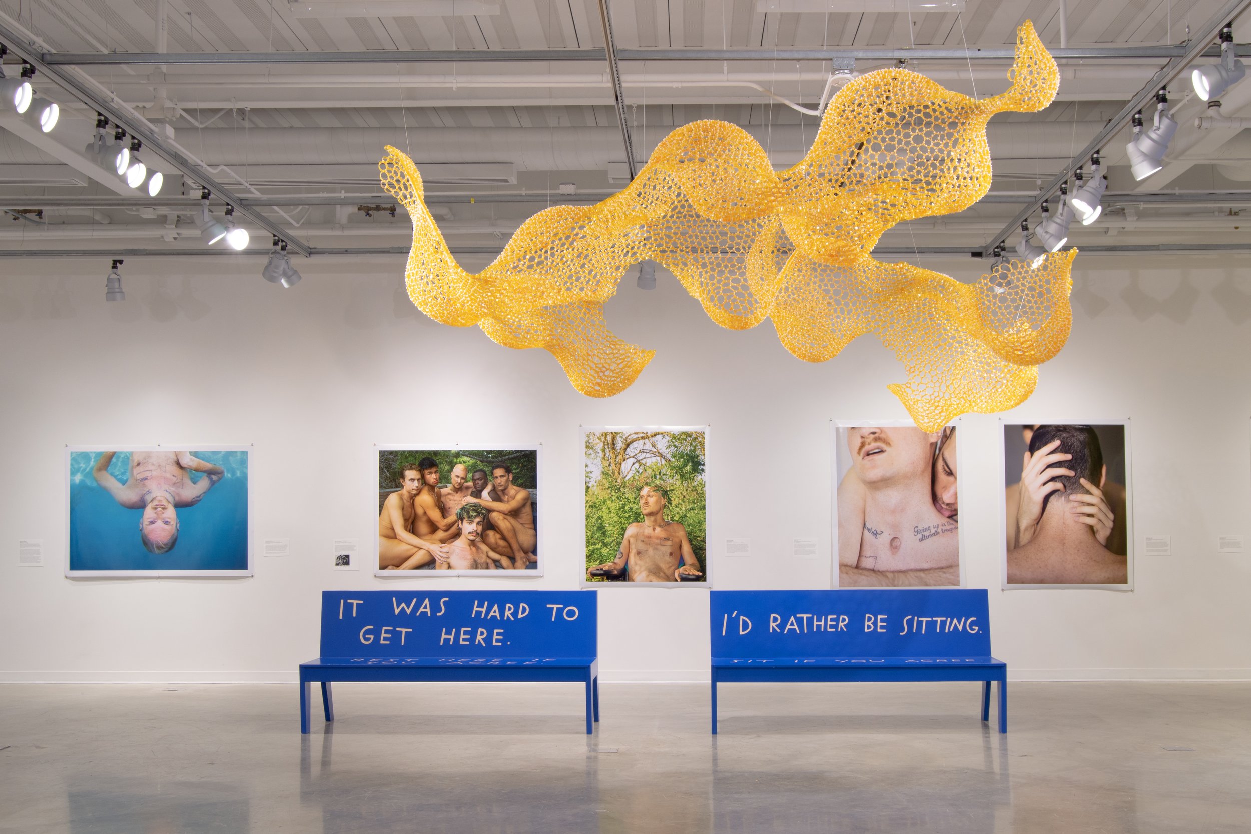 Art gallery with photographs of people and a yellow mesh sculpture hanging from the ceiling. Two blue benches with the words "It was hard to get here. I'd rather be sitting." in front of the wall.