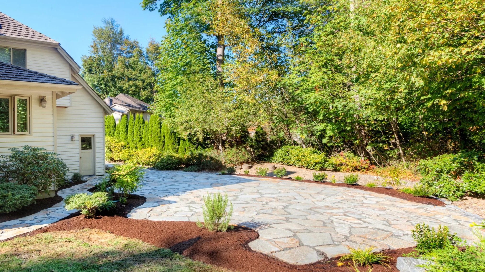 A landscaped backyard with a stone pathway, various green shrubs, plants, and tall trees in the background next to a light-colored house with a sloped dark roof.