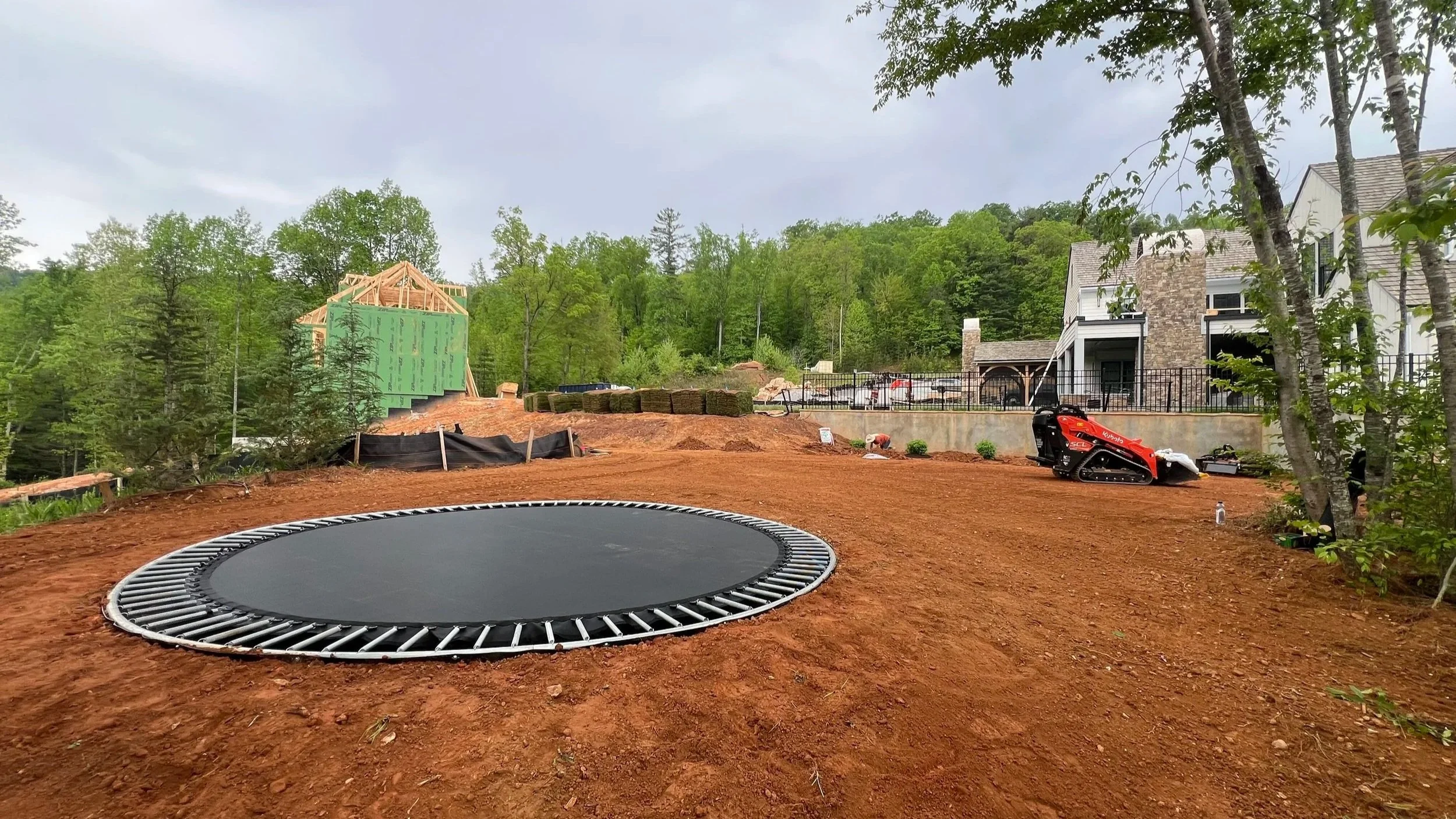 Construction site with a small trampoline in the foreground and a house under construction in the background, surrounded by trees and greenery.