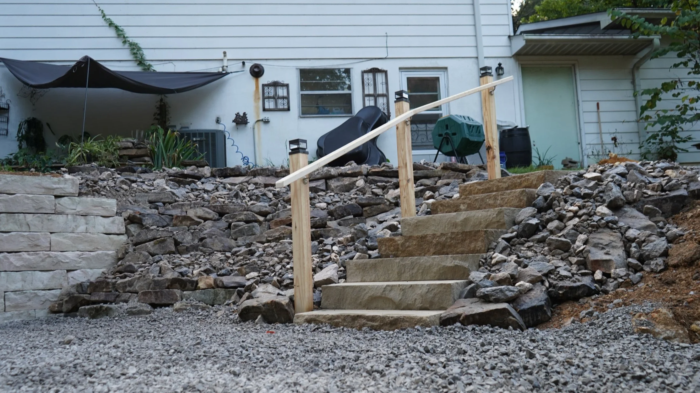 A set of stone stairs with a wooden handrail leading up a rocky slope in a backyard. In the background, there's a white house with windows, some garden tools, a black plastic chair, and a small covered patio area.