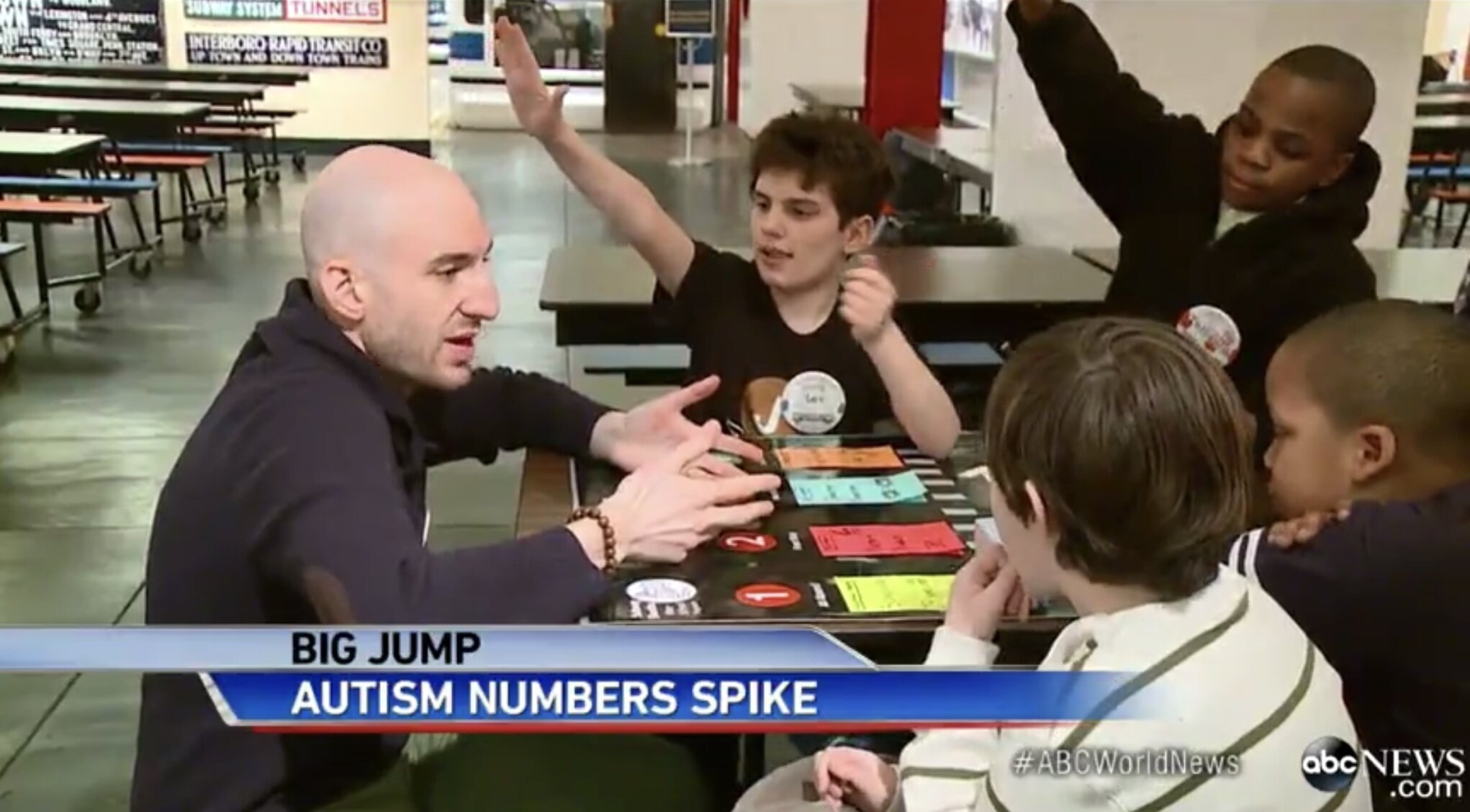 Aaron leans in front of a table in front of four elementary aged students, two of whom are raising their hands. A banner at the bottom reads "Big jump: autism numbers spike" with an "abc" logo in the corner.