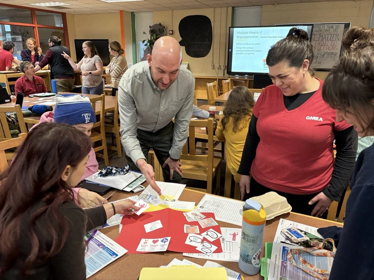 At a teacher workshop, Aaron points to materials on a table surrounded by teachers smiling and talking. In the background groups work at other tables.