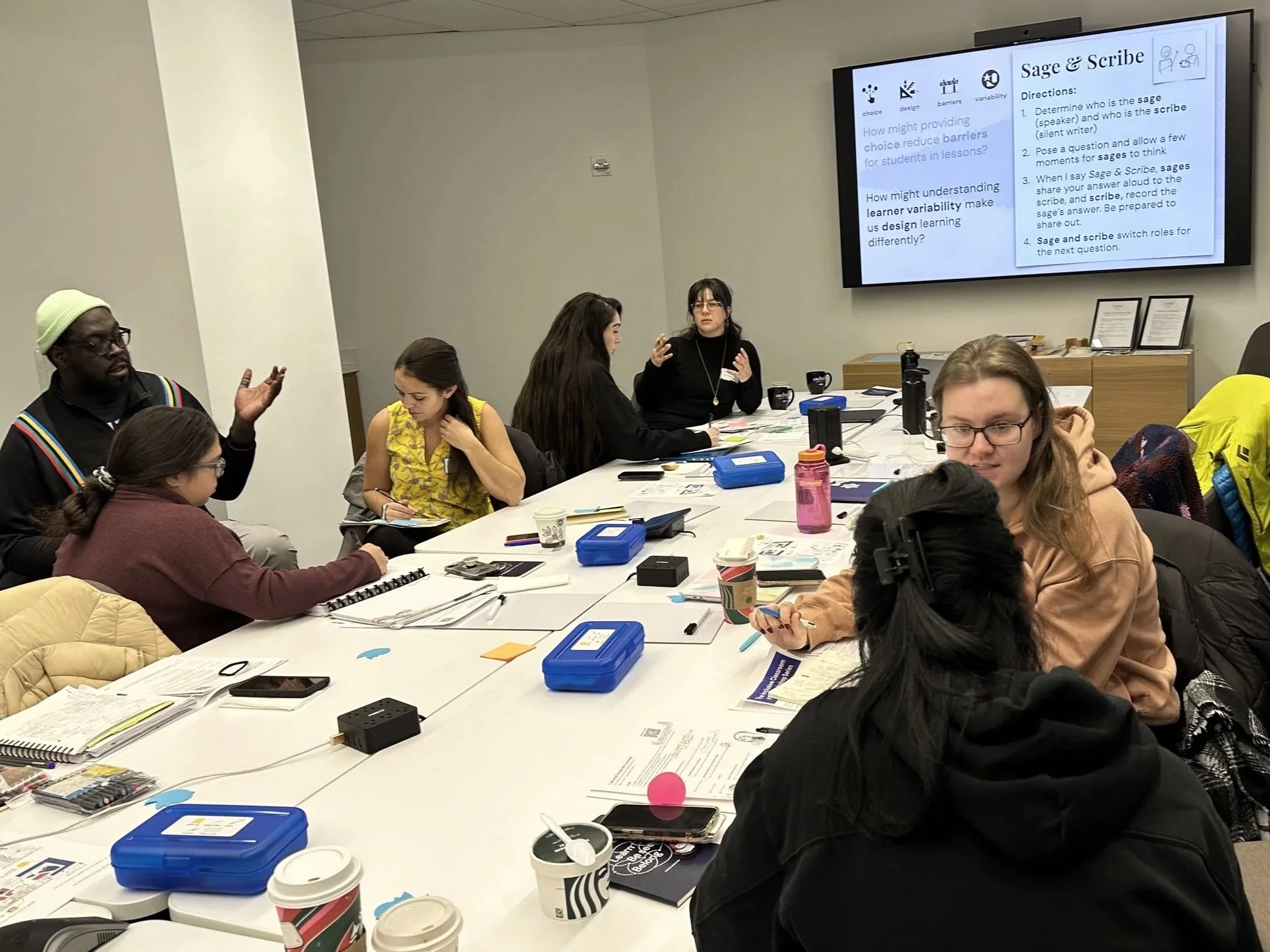 Group of educators sit around a long table covered in materials. They talk in groups of two. In the background are directions for an activity "Sage & Scribe."