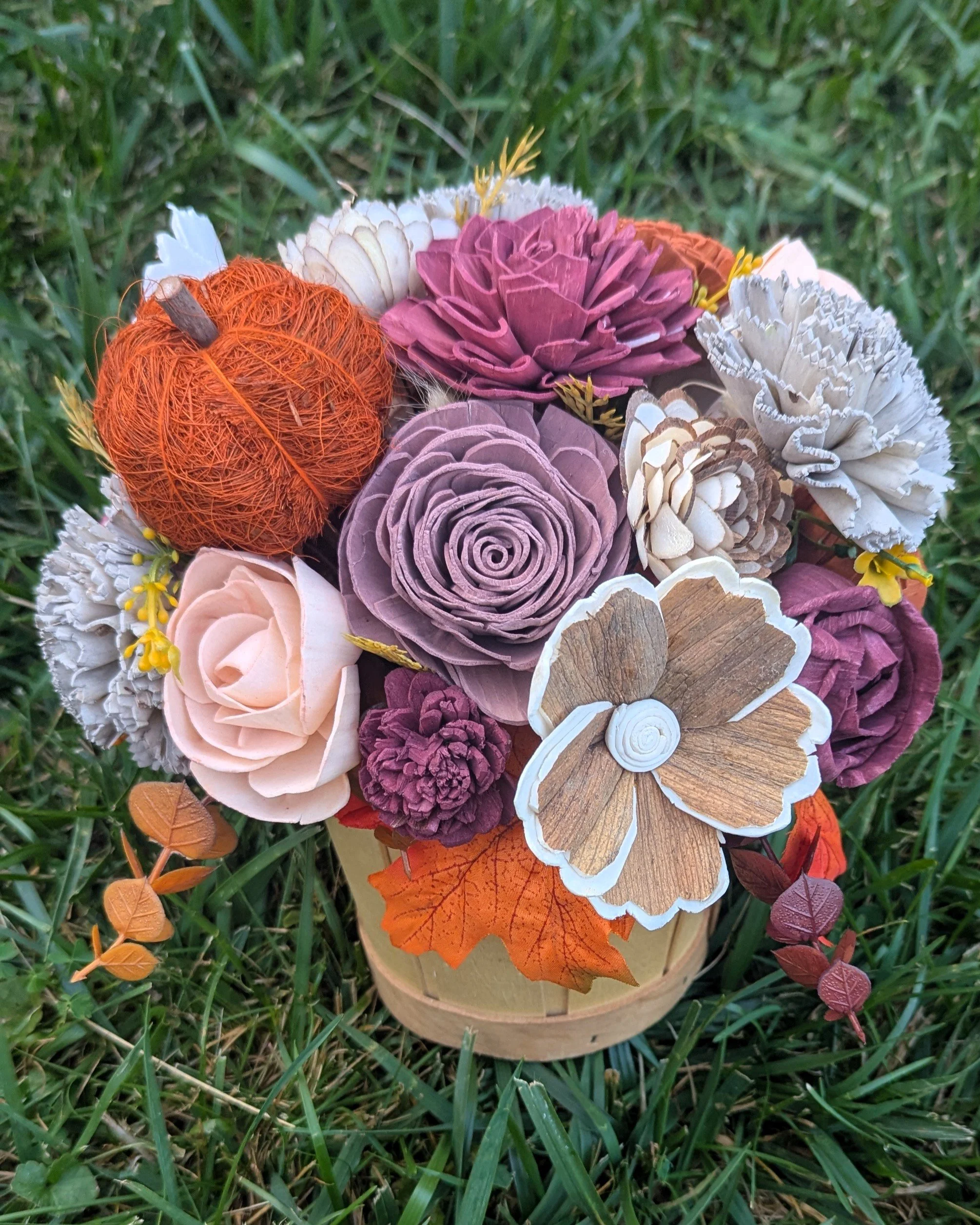 A sola wood flower arrangement of various autumnal colors, nestled in a wooden harvest basket.