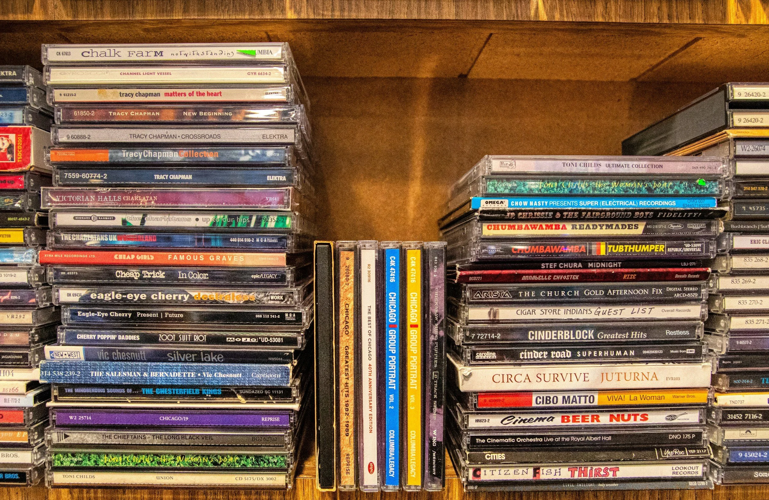 A large collection of CDs and cassette tapes stacked on wooden shelves, including various music albums and compilations.